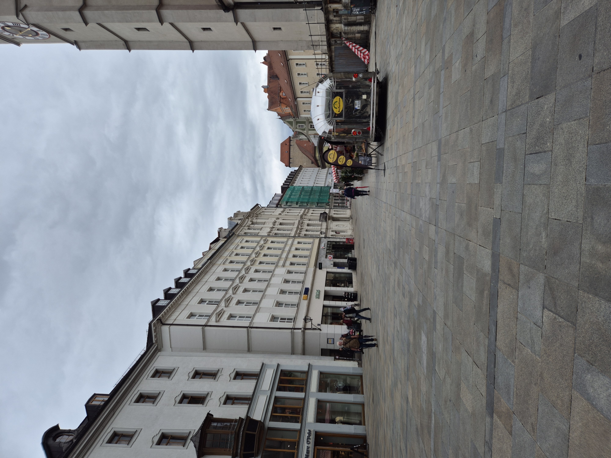 A street scene in Regensburg, Germany, captures a wide, paved plaza lined with historic buildings and a clock tower. The sky is overcast, and the architecture suggests a European city. The buildings on the left feature white facades with arched windows and traditional dormers. One building has "Marc O'Polo" signage, indicating a retail establishment. Further down the street, shops with glass storefronts are visible.
On the right side of the plaza, a prominent clock tower rises, a classic architectural element. Adjacent to it, a silver Airstream trailer, likely a food vendor, is parked. It has yellow signage with German text, possibly indicating its purpose as a bakery or food stand. Some people are present in the plaza, walking and interacting. One individual is walking away from the camera with a red umbrella, and another person is leaning against a railing. The paved ground is laid with large, square stone tiles. The scene conveys a sense of urban life on a cloudy day.