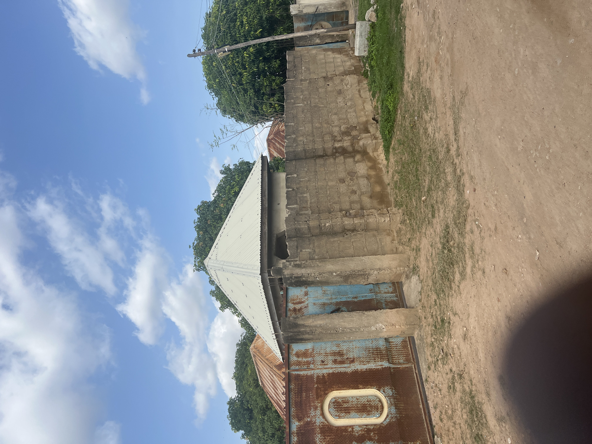 The image depicts a street scene in Jalingo, Nigeria, characterized by a weathered, rusty metal gate with an oval inset on the left. Adjacent to the gate is a light blue metal structure, likely another part of the gate or an entrance. A rough, cinder block wall, showing signs of wear and tear, extends to the right, partially obscuring two roofs of buildings behind it. One roof is corrugated and appears to be rusty brown, while the other, closer to the foreground, is a lighter, possibly galvanized metal, with a pyramidal shape. Lush green trees are visible behind the structures, and the sky above is a clear blue with scattered white clouds. The ground in the foreground is dusty and uneven, with patches of sparse grass. A wooden utility pole stands to the right of the wall, with wires extending from it. The overall impression is one of a typical residential or commercial area in a tropical climate, with elements of everyday life and the effects of time and weather visible.