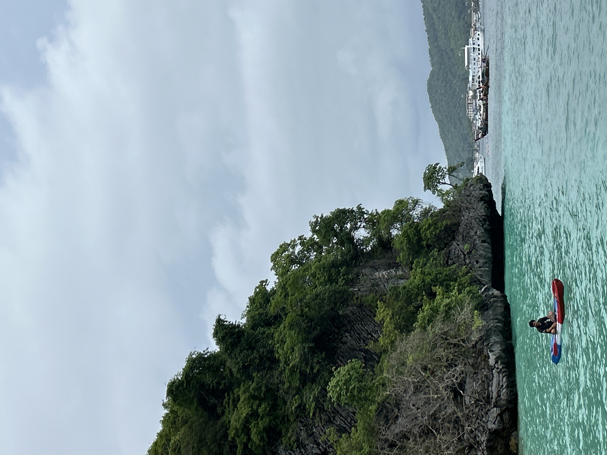 The image captures a view of Haad Hin Khom, Thailand. A rocky outcrop covered in lush green vegetation dominates the scene. Clear turquoise water is visible in the foreground, with a single person seated on a colorful inflatable raft or kayak. In the background, a coastline with buildings is visible against a cloudy sky. The photograph appears to be taken from a slightly elevated position.