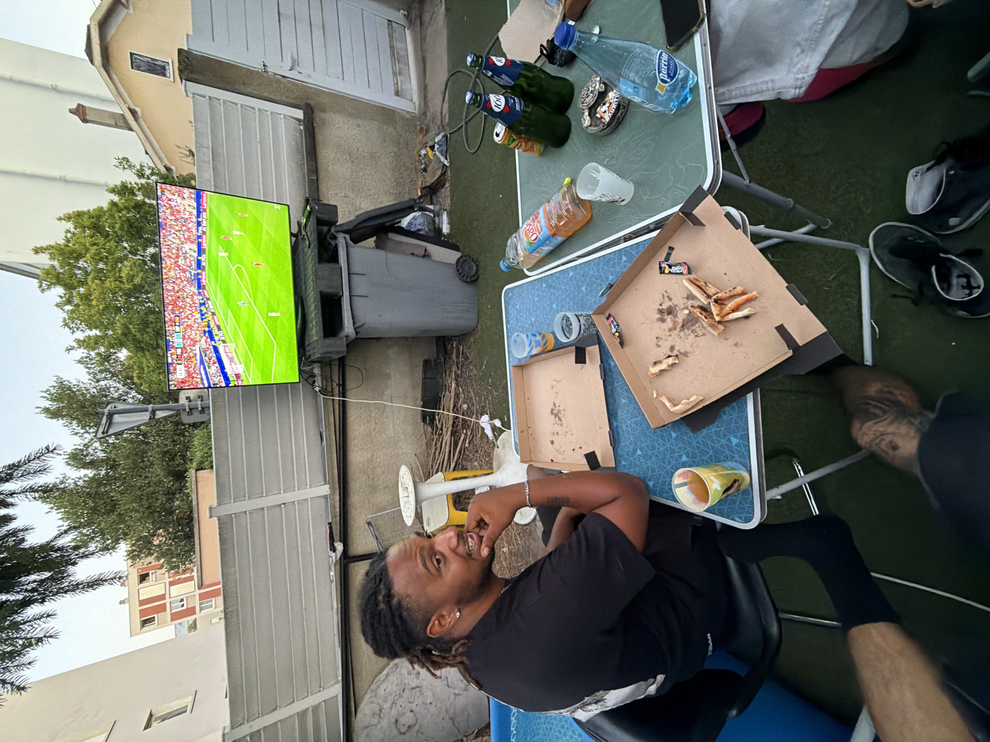 A man with dreadlocks, wearing a black t-shirt and bracelet, sits at a blue folding table in an outdoor backyard setting, looking to his right and holding his chin. A large flat-screen television displaying a soccer match is visible in the background, mounted on a gray plastic stand. On the table, there are empty pizza boxes, cups, a bottle of Oasis drink, two bottles of beer, and a bottle of Perrier. The ground is covered with artificial turf, and a blue inflatable pool is to the left of the man. In the background, residential buildings and trees are visible, suggesting an urban environment. The scene appears to be a casual gathering in Villeurbanne, France, with the man engaged in watching the soccer game while possibly having a meal and drinks.