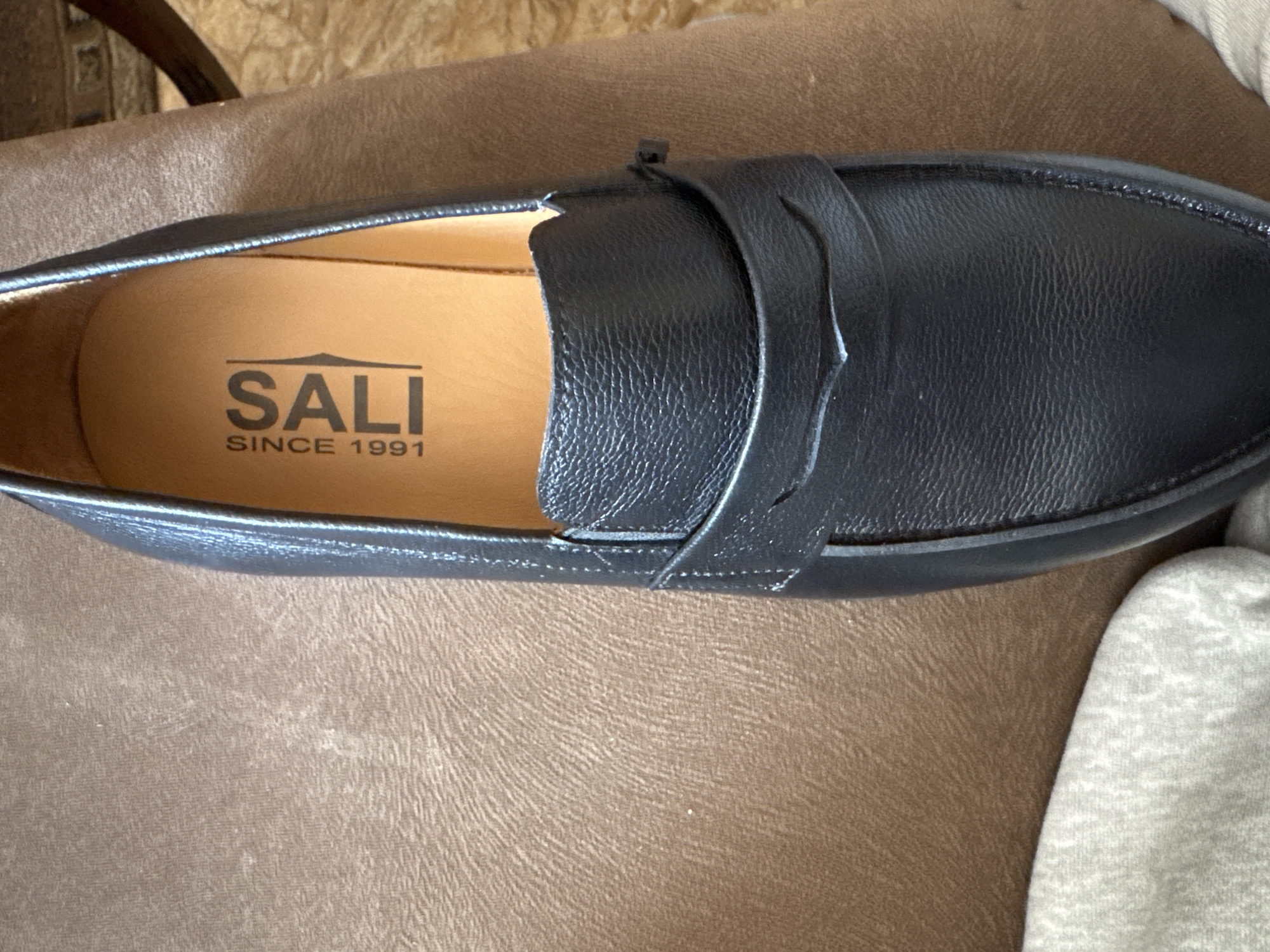 The image displays a close-up view of a single black leather loafer-style shoe, prominently positioned. The shoe features a textured, possibly pebbled, black leather upper with a classic penny loafer strap across the vamp. Delicate stitching is visible along the contours of the shoe and around the strap detail.
The interior of the shoe, specifically the footbed, is a lighter tan or light brown color. Clearly printed on the footbed is the brand name "SALI" in a dark, sans-serif font, accompanied by a small, stylized mountain peak graphic above the letter 'A'. Below the brand name, the text "SINCE 1991" indicates the brand's founding year.
The shoe rests on a textured, warm brown surface, likely upholstery such as a chair or couch. A soft, light grey fabric object, possibly an item of clothing, is partially visible and out of focus in the bottom right corner of the frame.
The scene depicts an indoor setting, with the shoe stationary, suggesting it is being displayed or photographed. There is no activity taking place, and no individuals are present. The image provides no specific visual cues to indicate the stated location context of Yerevan, Armenia, as the details are generic indoor elements.