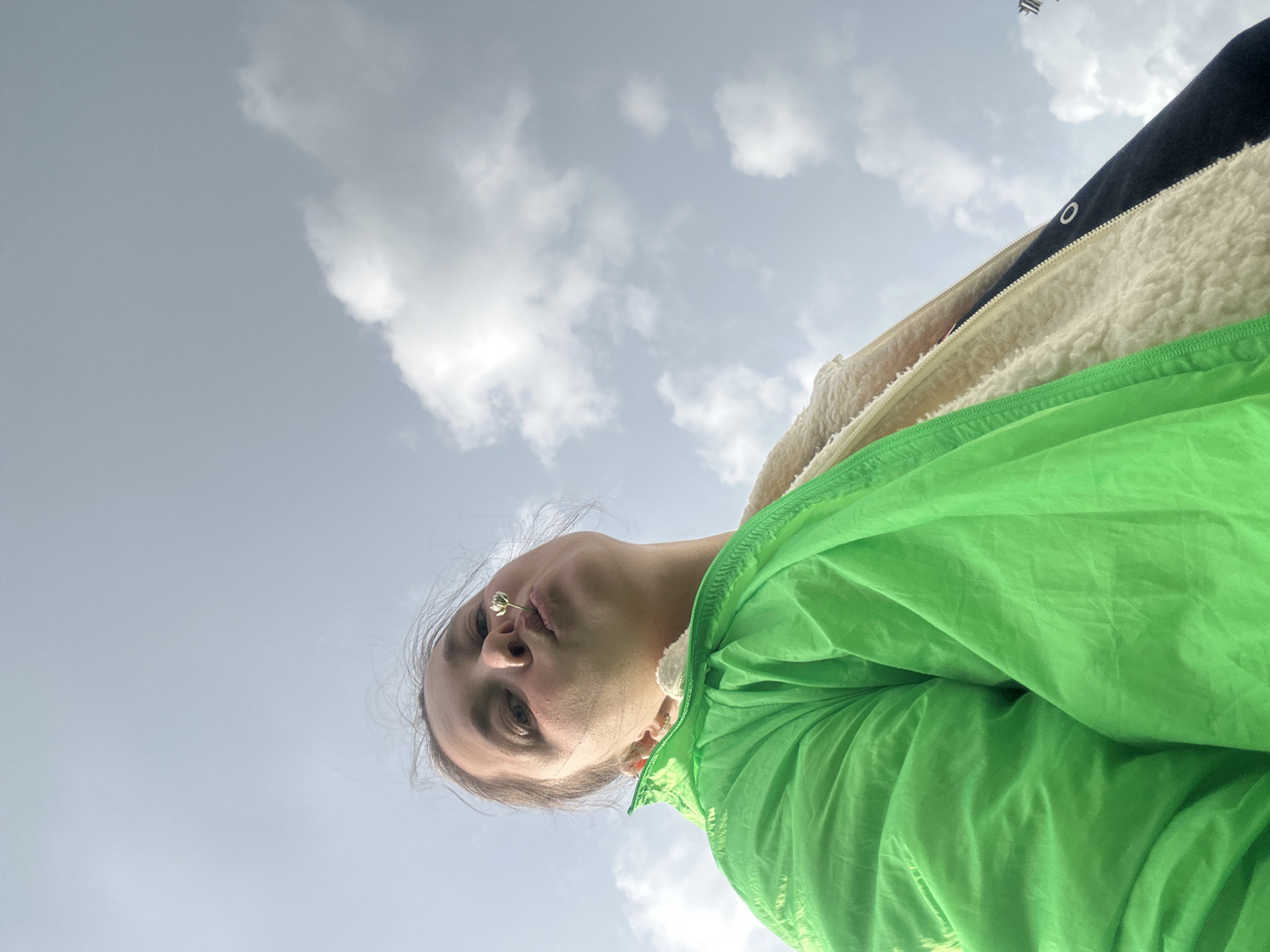 A low-angle, outdoor, chest-up selfie is captured against a cloudy sky. The subject, a young person, is wearing a bright green jacket over a cream-colored fuzzy interior layer and a black outer layer. A small white flower is positioned between their lips, suggesting they are puckering their mouth. The individual has light brown hair that is slightly windswept. The overall impression is casual and playful. The sky is filled with scattered white clouds. The location is Yaroslavka, Russia.