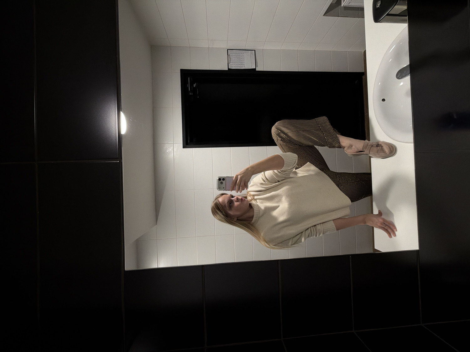 The image captures a blonde-haired woman taking a mirror selfie in a well-lit modern bathroom, likely in Izhevsk, Russia. She is reflected standing in front of a large mirror, holding a light-colored smartphone with both hands to take the picture. The woman is dressed in a cream-colored long-sleeved sweater, brown geometrically patterned wide-leg trousers, and light-colored loafers. Her expression is neutral as she focuses on her phone.
The bathroom features white square tiles on the walls. To the woman's right, a white oval sink with a metallic faucet is visible. Above the sink, a partially seen dispenser (possibly for paper towels or a hand dryer) and a small, framed piece of paper with illegible text are affixed to the tiled wall. A dark, likely black, door is reflected behind her in the mirror. The mirror itself seems to have a dark frame or is bordered by dark wall panels on the left and bottom edges, contributing to a clean and contemporary aesthetic. The setting is clearly indoors, with ample lighting.