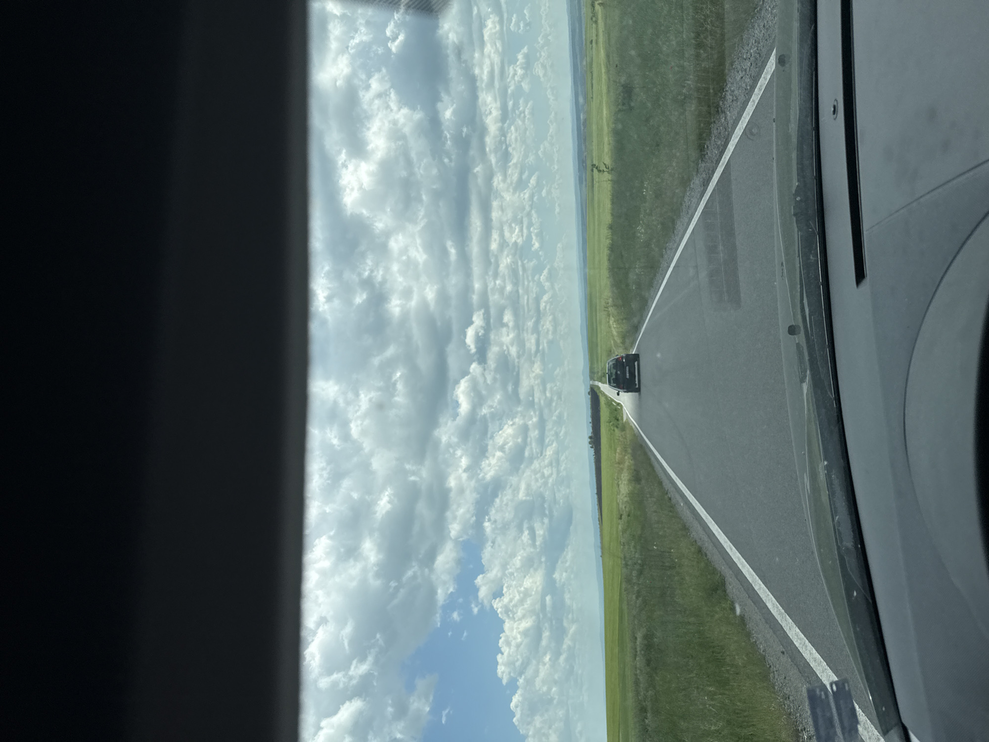The image shows a view from inside a vehicle, looking down a rural road. The road is paved with a white line marking the edge. Green grassy fields border both sides of the road. Another vehicle is traveling ahead in the same direction. The sky is visible on the left, filled with white and gray clouds. The vehicle's interior frame is visible on both the left and right sides of the image. The location is potentially in Troyanovo, Bulgaria.
