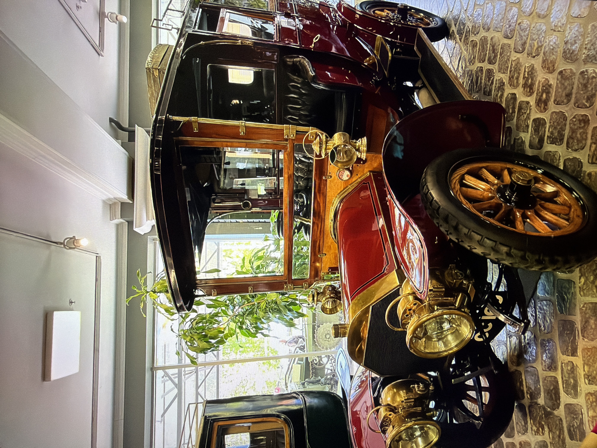 The image shows a vintage car displayed indoors. The car is a deep red with ornate gold accents, including the headlights, hood ornament, and trim. It has a dark, possibly leather, interior with wooden paneling. The car is parked on a cobblestone floor, and in the background, there is a large window with greenery visible outside. Part of another vehicle, painted black, is visible to the left of the main subject. The lighting suggests an indoor exhibition space or museum. The location in Kotelniki, Russia, is not visually discernible from the image. There are no people clearly visible interacting with the car.