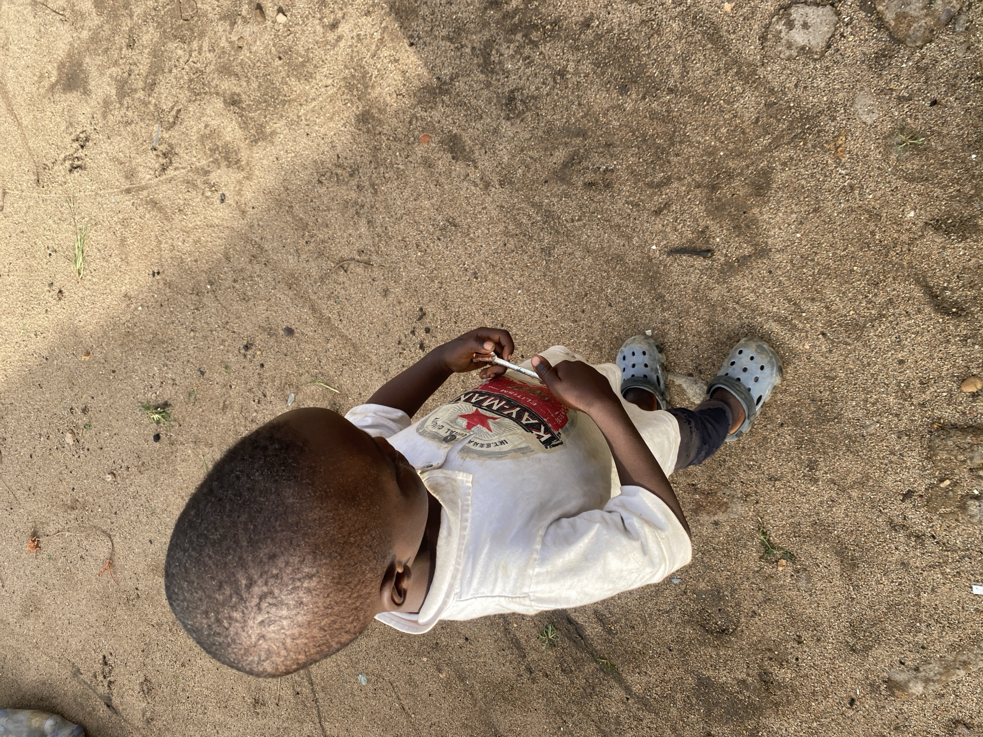 A child, appearing to be a young boy, is standing on a sandy surface. He is wearing a white polo shirt with a red and black logo, black leggings, and grey Crocs. He is holding a small object, possibly a syringe or pen, in his right hand. The ground is composed of loose sand, with some scattered small stones and a few blades of dry grass. In the background, the sandy ground continues with some indistinct shadows cast from overhead. The scene is outdoors, under natural light. The location is specified as the city of Jos, Nigeria.