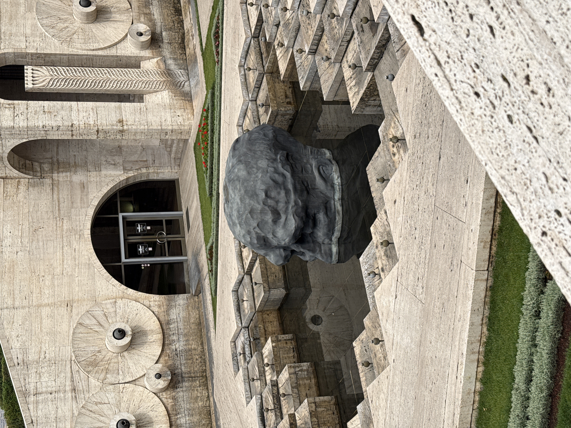 The image shows a section of the Cascade in Yerevan, Armenia. The architecture is constructed from light-colored stone. On the left is a multi-tiered structure with circular elements and an arched doorway. A large, dark, sculpted head sits on a tier of steps in the center of the image, flanked by narrow strips of grass and flowers. The angled perspective shows the intricate stepped design of the cascade, with small decorative elements along the edges. The overall scene is a mix of architectural details, landscaping, and sculptural art.