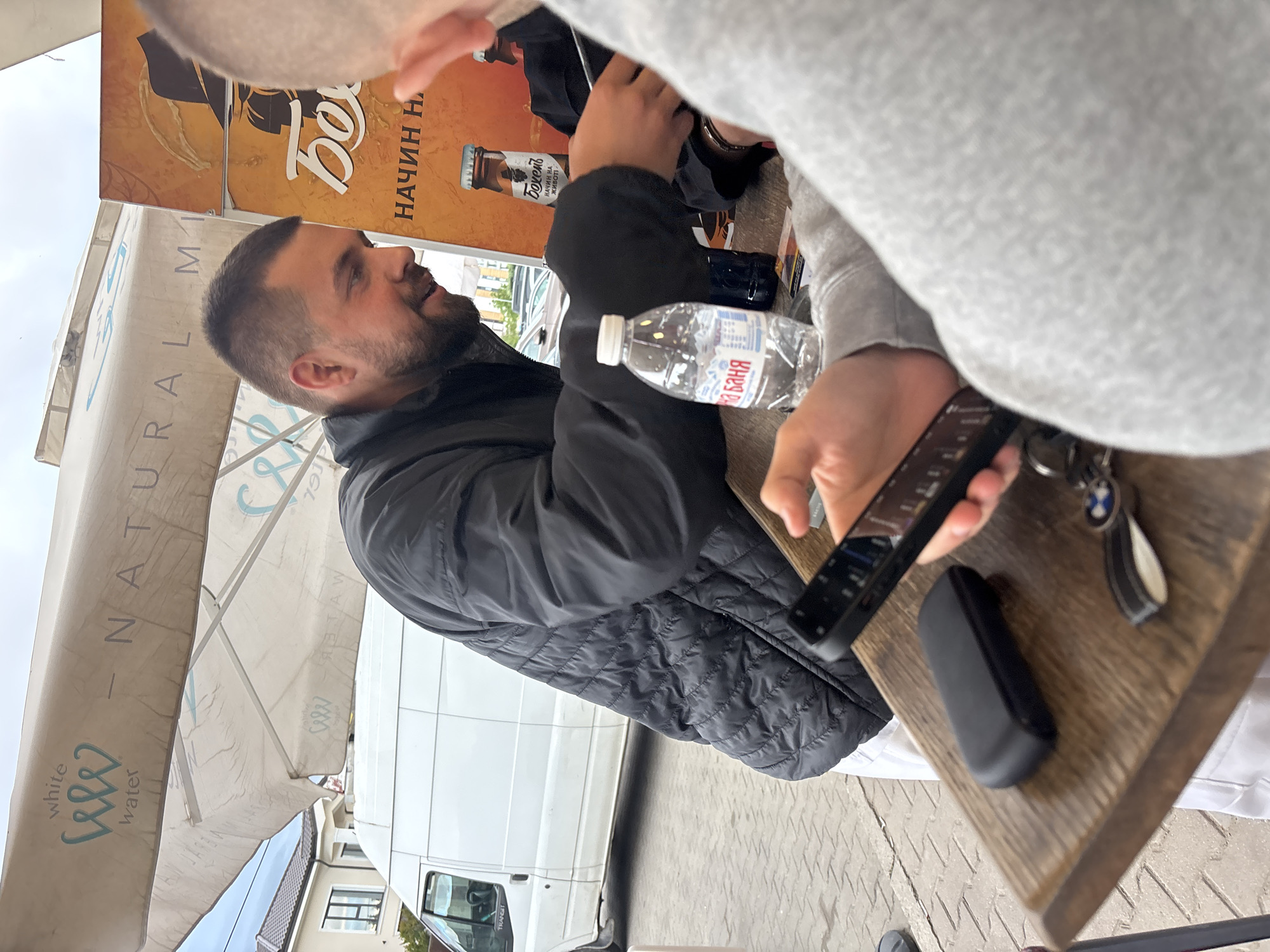 Here's a forensic description of the image:
**People:** Two men are visible. One man is in focus and has short dark hair and a beard, wearing a black quilted jacket. Another man is partially visible, wearing a grey sweatshirt.
**Objects:**
* A water bottle labeled "Баня" (Banya).
* A smartphone.
* A pair of keys, with a BMW logo visible.
* A black, oblong object.
* A table with a wooden surface.
* An umbrella with "NATURAL MI - white water" printed on it.
* A sign advertising "Бохемъ" beer.
* A white van.
**Actions/Interactions:** One of the men is holding the smartphone.
**Environment:** The scene appears to be an outdoor setting, possibly a cafe or restaurant. There is a van parked nearby. The writing on the sign and water bottle suggests the location is Bulgaria. The presence of "Бохемъ" (Bohemian) beer may indicate the location is near Plovdiv, Bulgaria.