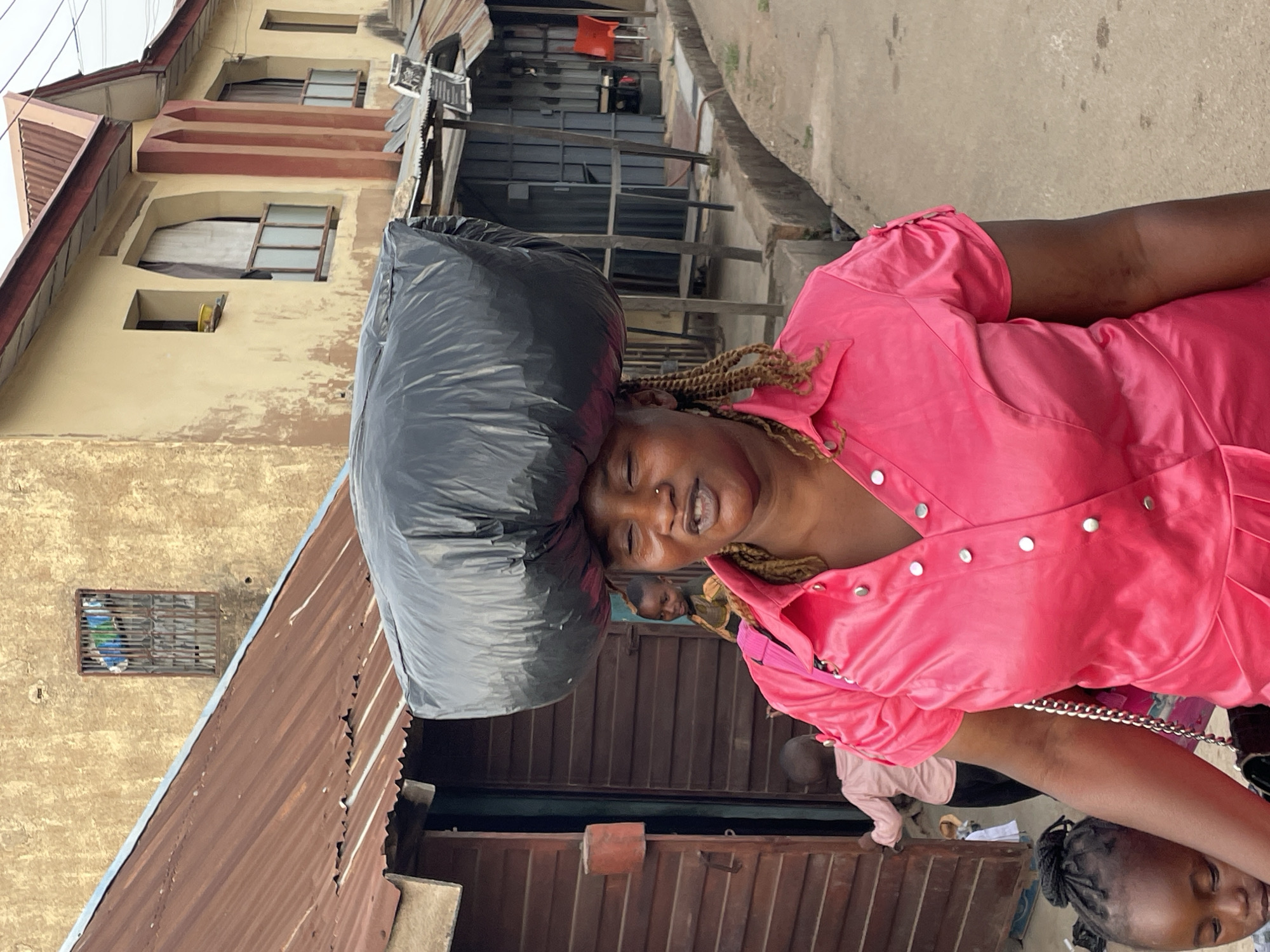 The original image, rotated 90 degrees clockwise, captures an outdoor urban scene in Jos, Nigeria. It features a woman balancing a large, dark grey or black plastic bag on her head. The woman, positioned centrally, wears a vibrant pink top or dress adorned with silver buttons down the front. She has braided hair, a nose piercing, and is smiling broadly with teeth showing, looking towards the viewer.
In the lower right foreground, a child's face is partially visible. Further back, another person's head is indistinctly seen behind the main woman. In the deeper background, to the right, a person in a light-colored top is visible near a structure.
The setting includes a multi-story building with a light-colored (tan or beige) facade and brown architectural elements on the left side of the frame. Several windows, some with visible wooden frames or coverings, are present on this building. Below it, a structure with corrugated reddish-brown metal roofing is visible. The background also shows indistinct buildings or market stalls with dark metal structures and gates. A concrete curb or path runs along the right side of the frame.
The photograph appears to be taken during daylight hours, under bright conditions. The woman's cheerful expression is notable. No discernible text is visible within the frame. The act of carrying goods on her head is a common practice in the region.