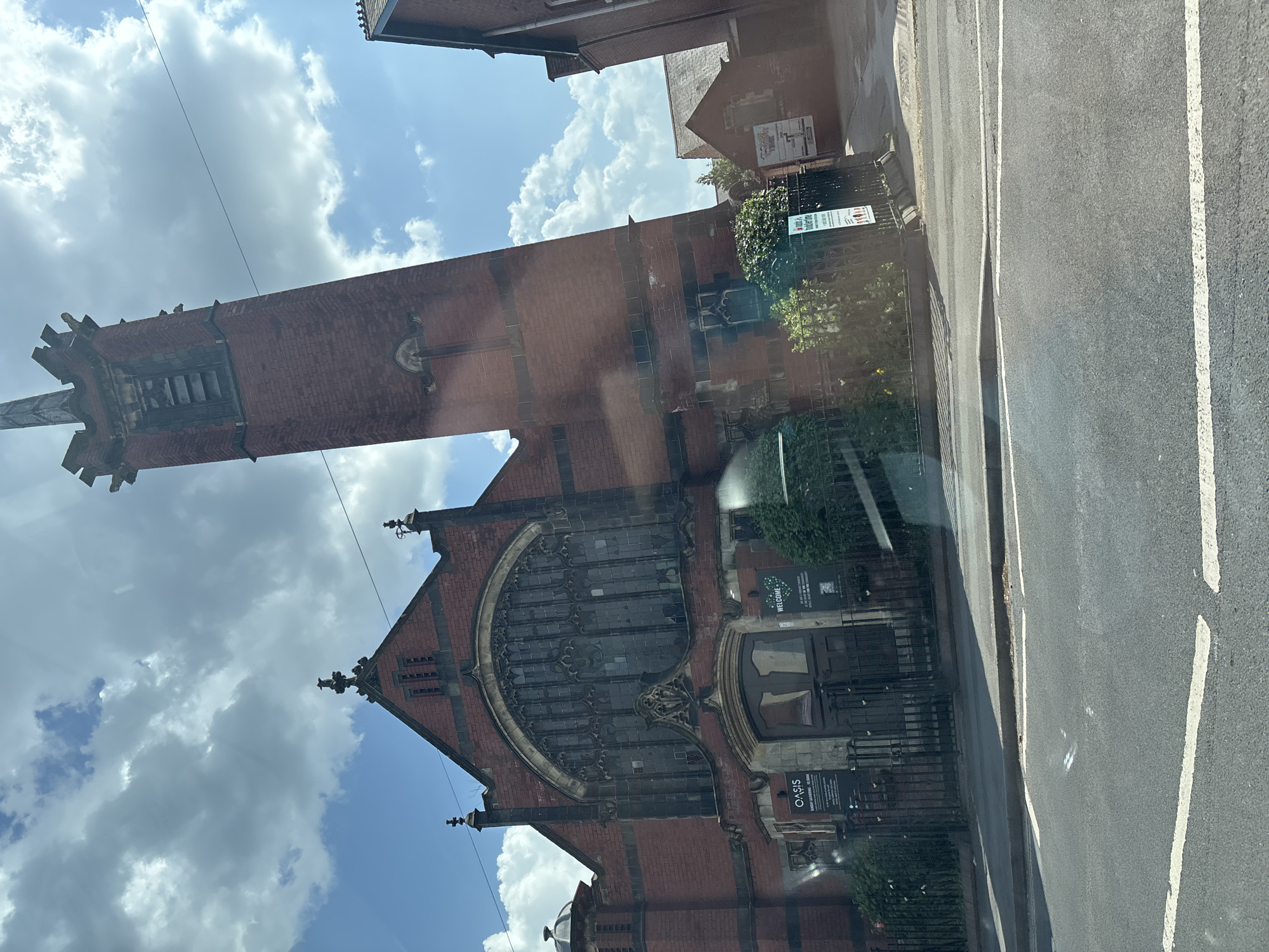 The image shows a street-level view of the exterior of the building, the Oasis Nottingham. A tall, red-brick tower with a detailed apex stands prominently against a blue sky filled with fluffy white clouds. The front facade of the building features a large arched window with intricate stone tracery. Black metal gates and fencing run along the perimeter of the property. In the foreground, a street with white lane markings is visible, suggesting the scene is taken from inside a vehicle. The building appears to be in Nottingham, United Kingdom, judging by the name of the building.
