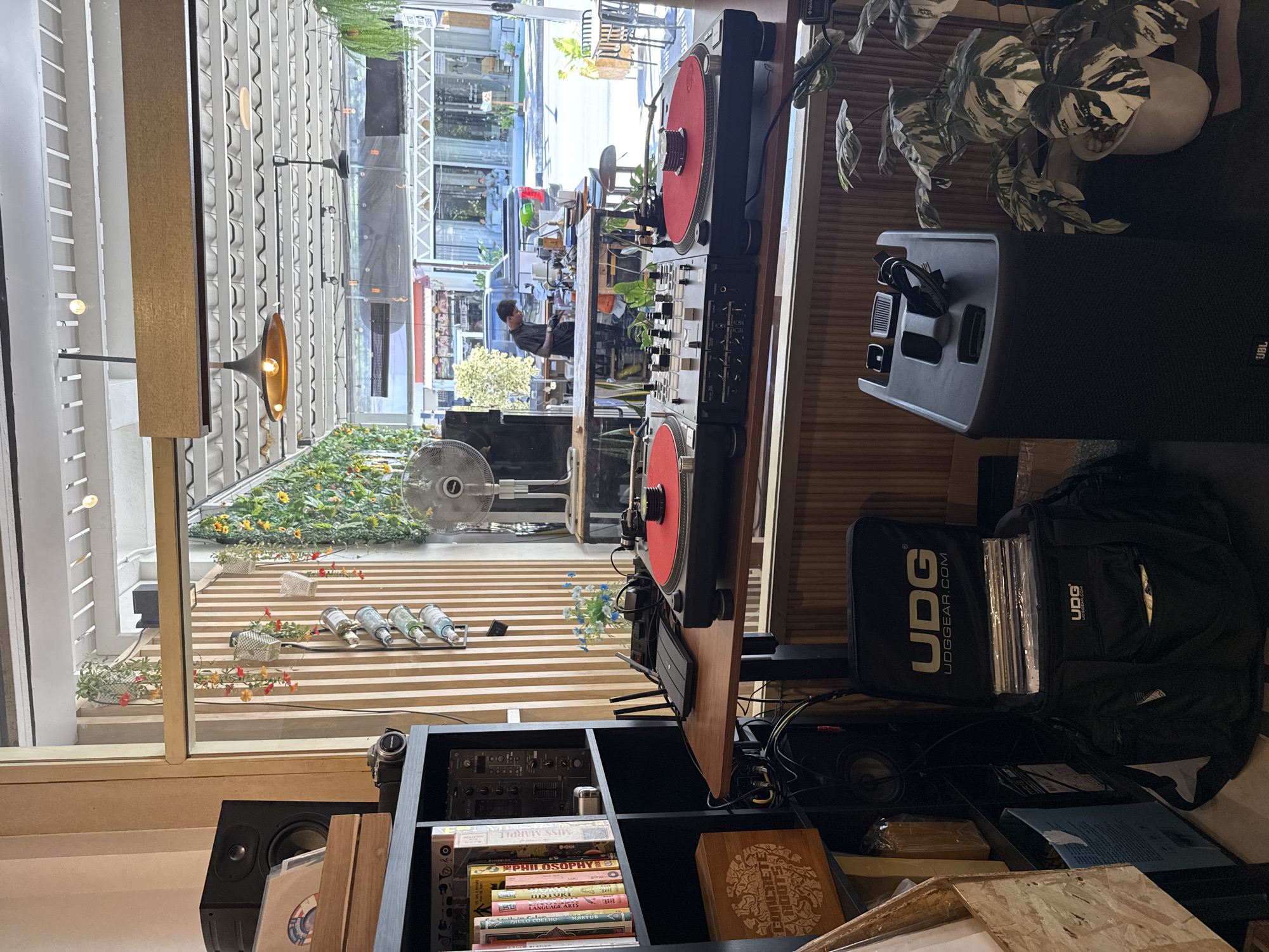 The image depicts an indoor scene, likely a cafe or bar, in Koh Samui District, Thailand. The foreground features a DJ setup with two turntables and a mixer positioned on a wooden counter. A large black speaker stands to the right, and a UDG branded bag filled with records is on the floor to the left. A bookshelf filled with books and other items is also visible on the left. Through a large window, the exterior of the establishment can be seen, including street activity and other shops. The overall ambiance suggests a place that combines music and social gathering.