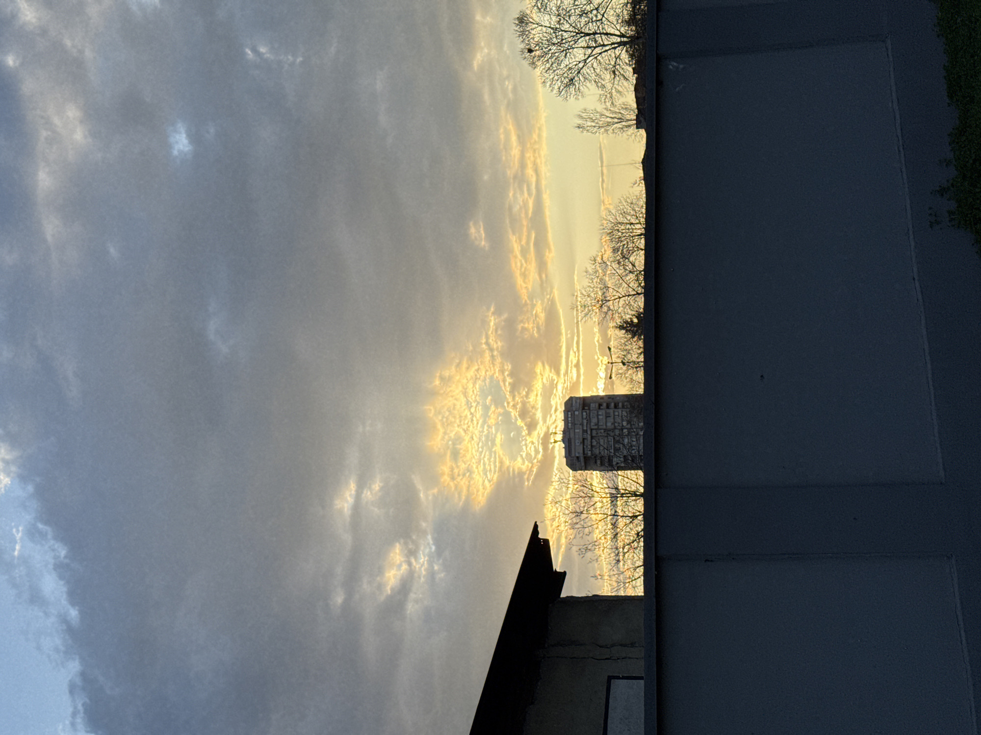 The image shows a view towards the sky at sunset in Plovdiv, Bulgaria, obscured by buildings and trees. The sky is filled with gray clouds, with some streaks of yellow light shining through. The silhouette of a multistory building with windows is visible, as well as trees. Other buildings are partially visible at the bottom of the frame. A dark gray surface occupies the right side of the frame, possibly a wall or street.