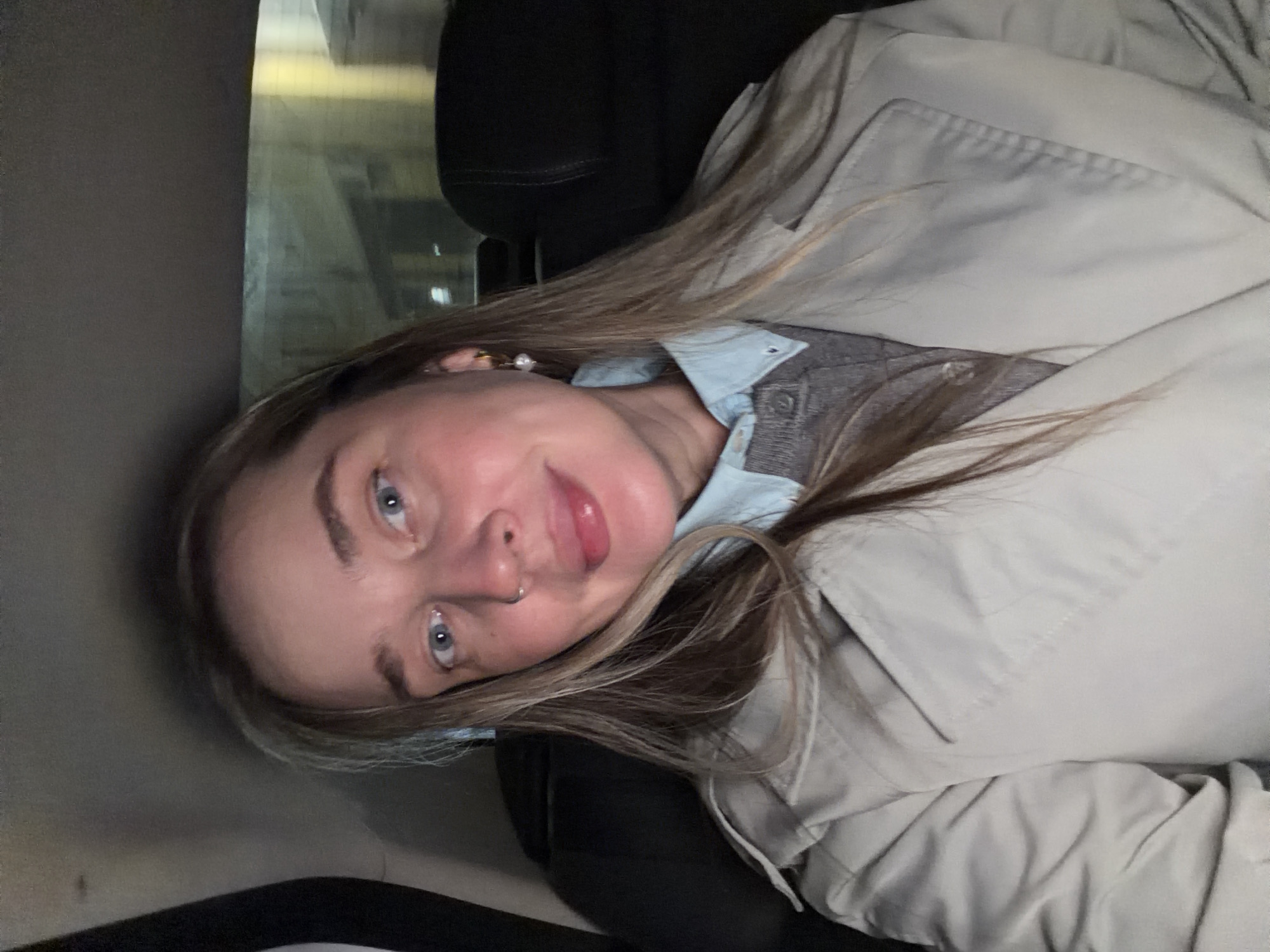 A young woman with light skin, blue eyes, and blonde hair is the main subject of the photograph. She is wearing a light-colored trench coat over a gray vest and a light blue button-down shirt. The woman is wearing a nose ring and earrings. The background shows what appears to be the interior of a vehicle. The specific location in Moscow, Russia, cannot be determined from this image.