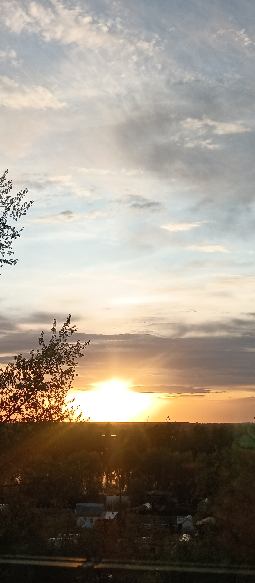 The image captures a sunset over a landscape featuring trees, buildings, and cranes. The sun is positioned near the horizon, emitting a bright light. Clouds are visible in the sky above. The location appears to be a residential area with industrial elements.