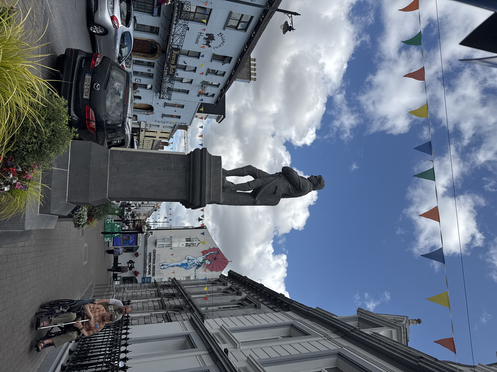 In Clonmel, Ireland, a statue of a man stands on a pedestal in a town square. The statue is made of dark stone and depicts a man with his arms crossed. He wears clothing that appears to be from the 18th or 19th century. Behind the statue, a row of buildings lines the street, including a light blue building with "Bianconi House" and "Keatons Hotel" signage. Colorful triangular flags are strung across the street, decorating the scene. A black Mazda MPV with license plate "132-D-5764" is parked in the street. In the foreground, an elderly man in a wheelchair is being pushed by another man. The scene appears to be a typical day in a European town, with people going about their business.