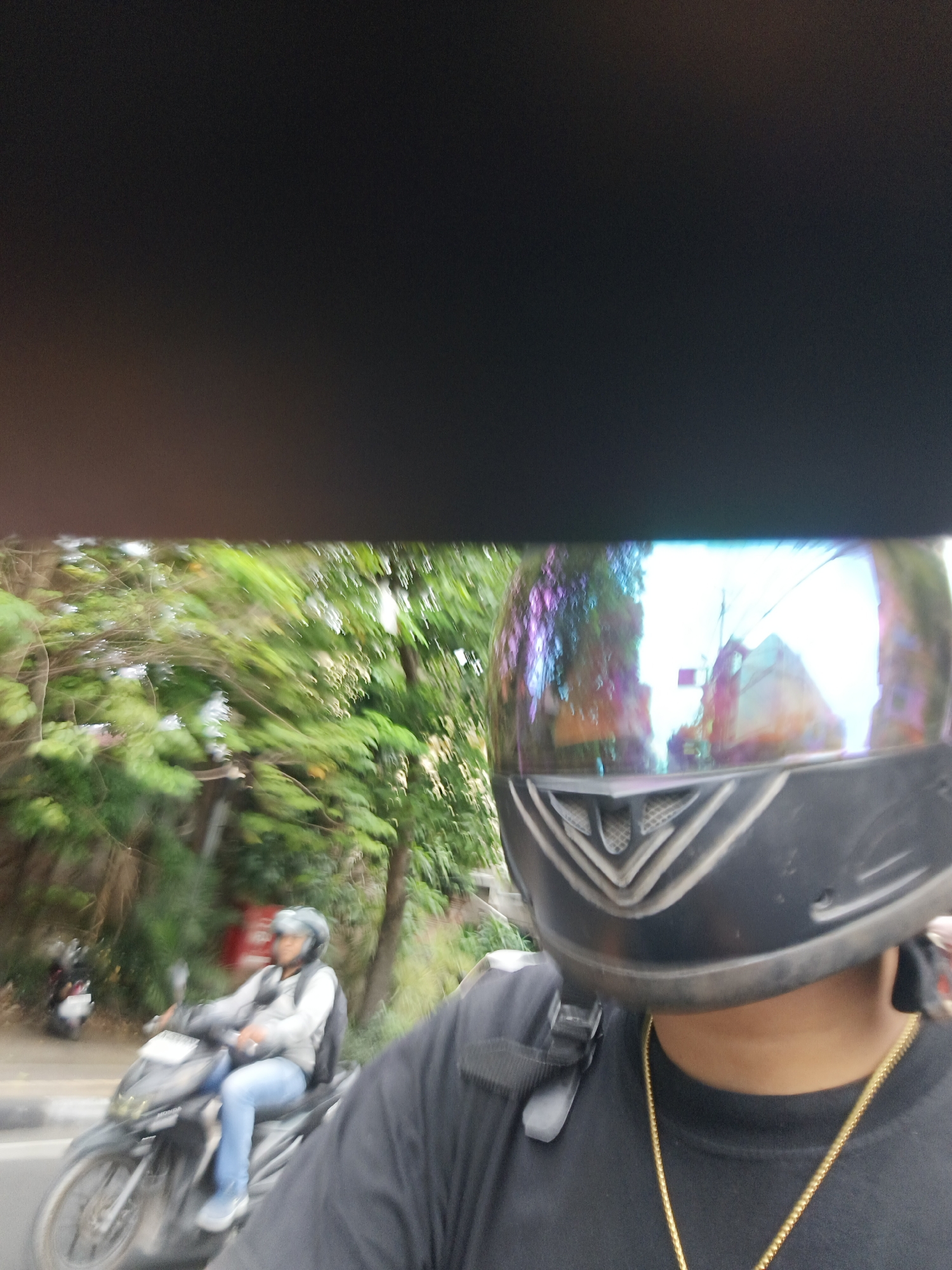 The image shows a first-person perspective of someone riding a motorcycle. The rider is wearing a black helmet with a reflective visor that shows a distorted, colorful reflection of the surroundings. The rider is also wearing a black t-shirt and a gold chain. In the background, another person is riding a motorcycle and appears to be on the left side of the road. The environment is lush with green trees and foliage, suggesting an outdoor setting in a tropical climate. The motion blur indicates that the motorcycles are in motion. The scene is likely from a street in Denpasar, Indonesia, given the common use of motorcycles for transportation in the city and the tropical vegetation.