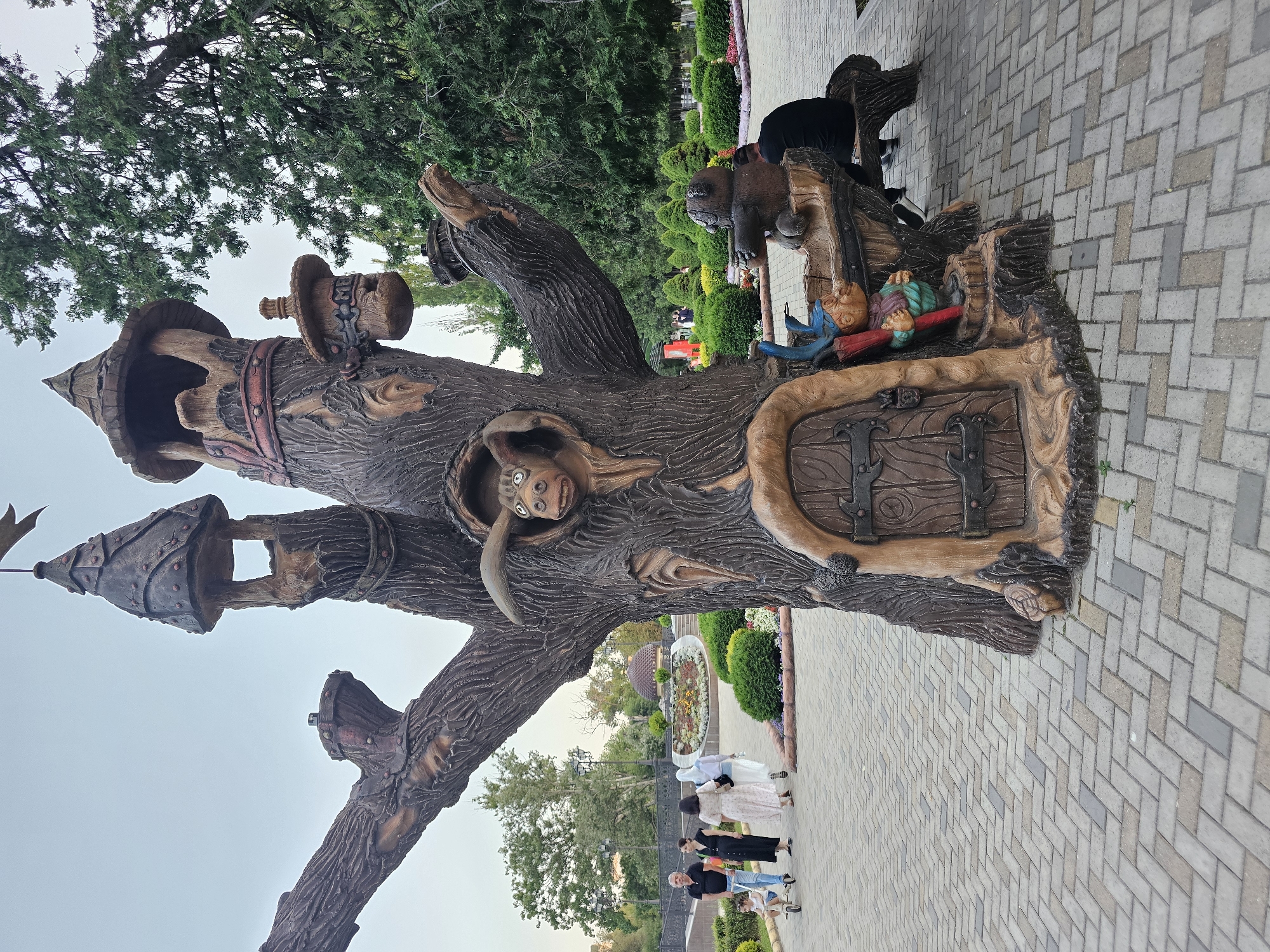 The image shows a decorative tree sculpture with fairy tale elements in a park. The sculpture is brown and textured to resemble a tree trunk with branches. It features a small door at the base, a carved face of an animal looking out from a hole in the trunk, and several tower-like structures on its upper branches. A gnome-like figure holding an umbrella is positioned near the door, and a bear sculpture is seated on a bench attached to the tree. The park setting includes a paved walkway with a herringbone pattern, trimmed green bushes, flowerbeds with colorful flowers, and tall trees in the background. Several people are walking in the park: a man with a child in a wheeled cart, and two women walking behind them. The sky is overcast. This sculpture is located in the city of Cherkessk, Russia.