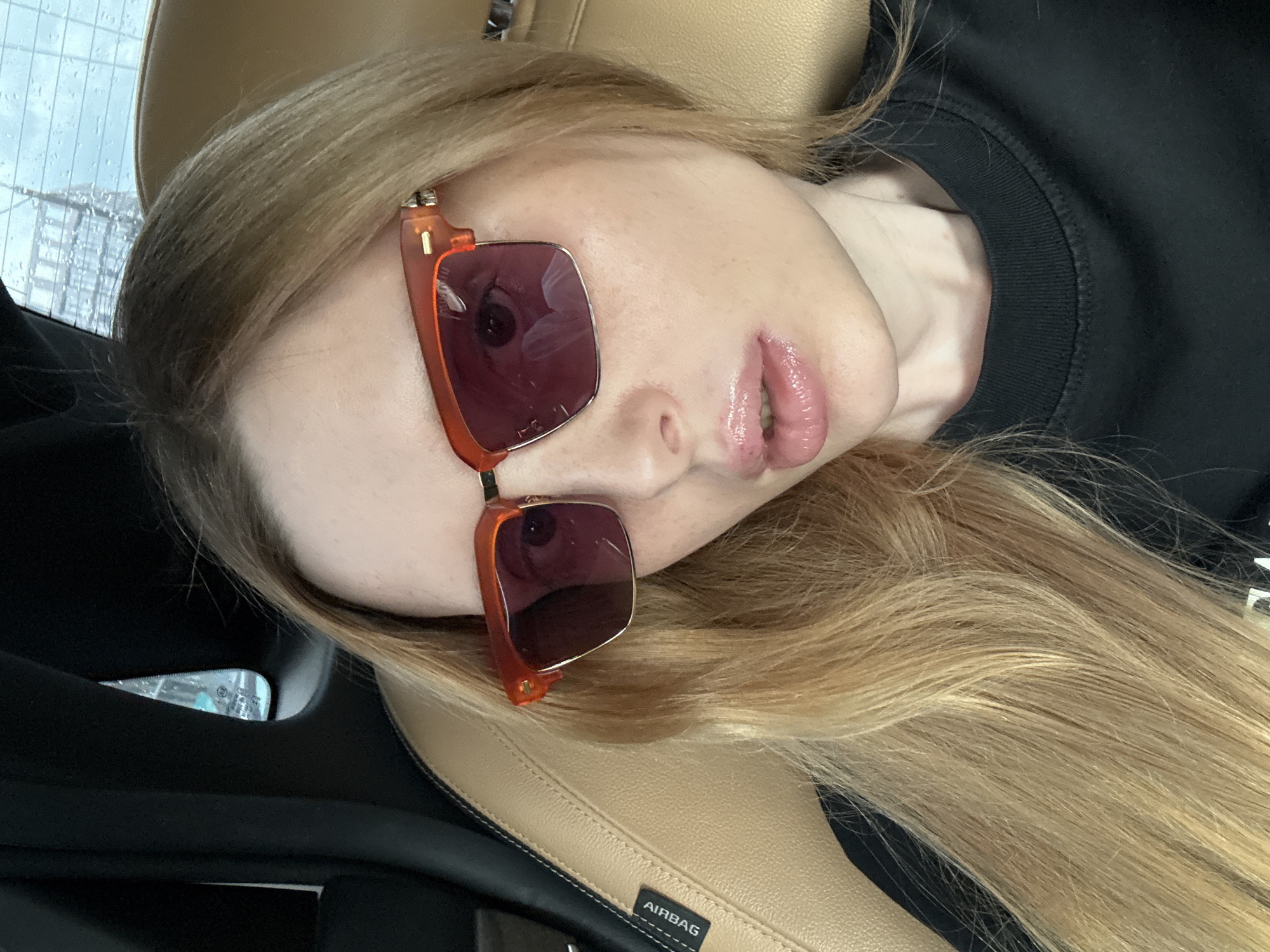 This is a close-up, high-angle shot of a young woman lying down, possibly in a car. She has long, blonde hair that falls around her face and shoulders. She is wearing oversized, square-shaped sunglasses with bright orange frames and dark lenses, which reflect a distorted image of what appears to be her eye. Her lips are parted, revealing slightly glossy, pink lipstick. She is wearing a black crew-neck sweatshirt. The background suggests an indoor setting, with the tan leather of a car seat visible in the foreground. There is a slight sheen on her skin, and the lighting is soft, hinting at natural light. The overall mood appears relaxed and casual. A small black tag with the word "AIRBAG" in white capital letters is visible on the car seat. The image is rotated 90 degrees counterclockwise.