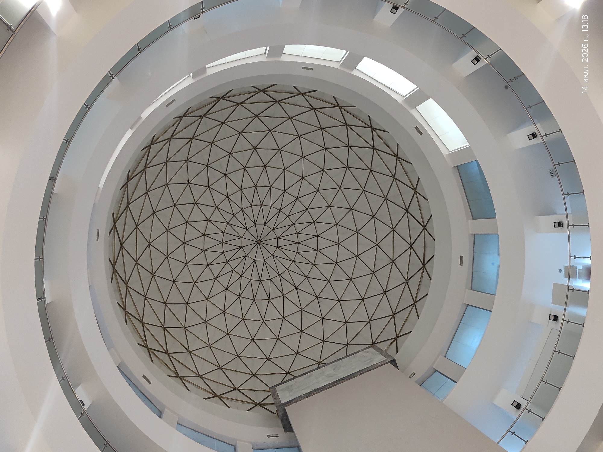 The image shows the interior of a domed building with a geometric ceiling. The dome is supported by a network of triangular golden-colored metal struts, creating a tessellated pattern. Surrounding the dome, there are multiple levels of curved balconies or walkways, made of white material and featuring glass railings with metal supports. Rectangular windows are visible on the lower levels, some of which appear to be tinted blue. There are also black spotlights mounted on the walls of the lower levels. The lighting suggests an indoor environment with natural light coming through the windows and artificial light from the spotlights. The overall aesthetic is modern and architectural, with a focus on geometric forms and clean lines. A timestamp in the lower right corner reads "14 июл. 2026 г., 13:18".