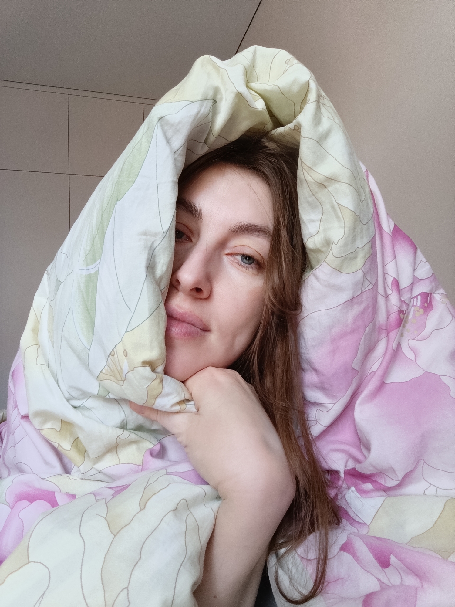 A woman with light skin and auburn hair is partially covered by a floral patterned duvet, with the duvet draped over her head, forming a hood-like shape. Her left eye is visible, and she rests her face on her left hand. The duvet has a pattern of pink and white flowers, and some yellow and green accents. The background shows a neutral-colored wall and a cabinet with a grid of rectangular sections. The image appears to be taken indoors, likely in a residential setting. The location in the city of Chaikovsky, Russia, is not discernible from the visual information provided.