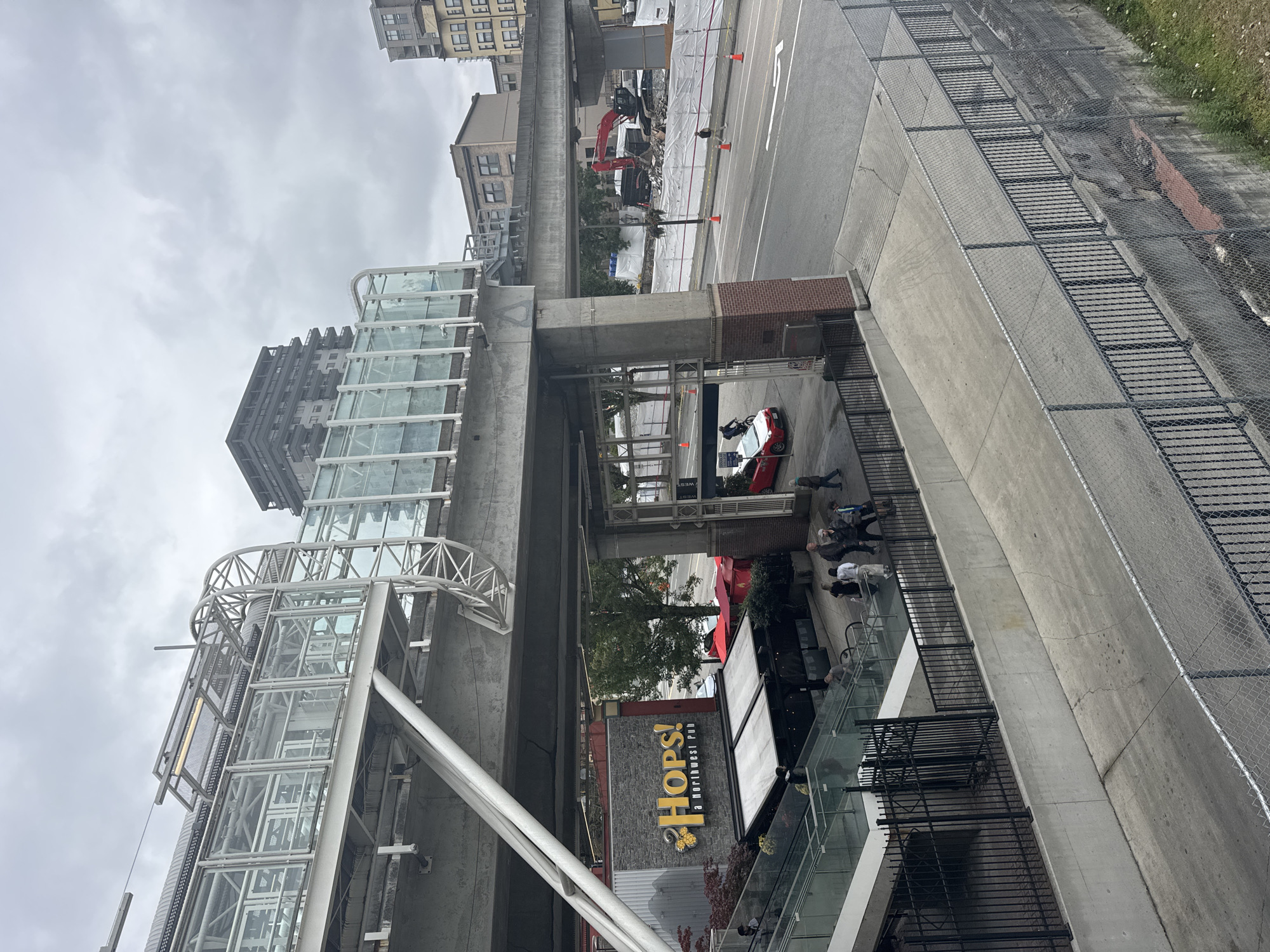 This image depicts an exterior view of a transit station and surrounding urban environment in New Westminster, Canada. The primary subject is an elevated, modern transit platform constructed with concrete and extensive glass paneling, supported by substantial concrete pillars. Below the platform, the facade of a building with a prominent "Hops! a Northwest Pub" sign is visible, featuring red awnings and glass storefronts.
Several individuals are present: a group walking on an escalator leading down from the platform, a person on a bicycle near a red car, and a few other pedestrians on the sidewalk and street level. The environment includes a street with traffic cones, construction barriers, and heavy machinery (a red excavator) indicating ongoing work in the background. Other buildings, including a tall, dark-colored apartment complex and multi-story commercial buildings, are visible in the distance. The sky is overcast, suggesting a cloudy day. The scene is captured from an elevated vantage point, looking down and across the transit infrastructure and street.