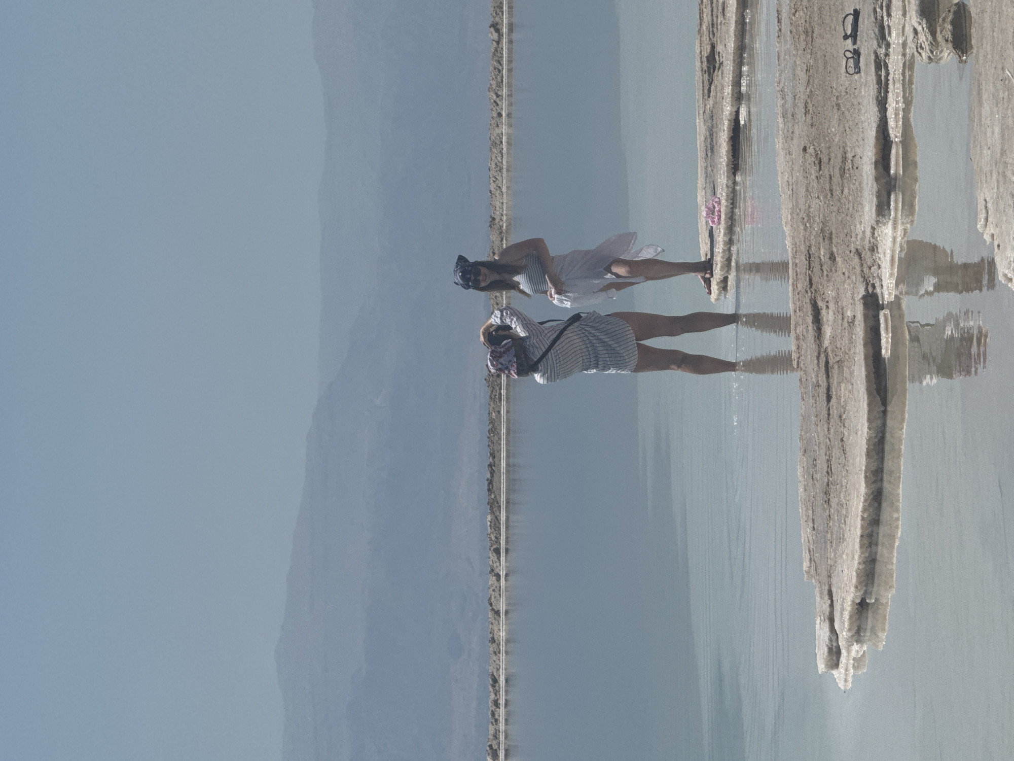 The image shows two women standing in the Dead Sea, in the city of Tamar Regional Council, Israel. One woman is wearing a white dress and a mask, while the other is wearing a striped shirt and shorts. Both women are standing near salt formations in the water. In the background, there is a mountain range and a clear sky.