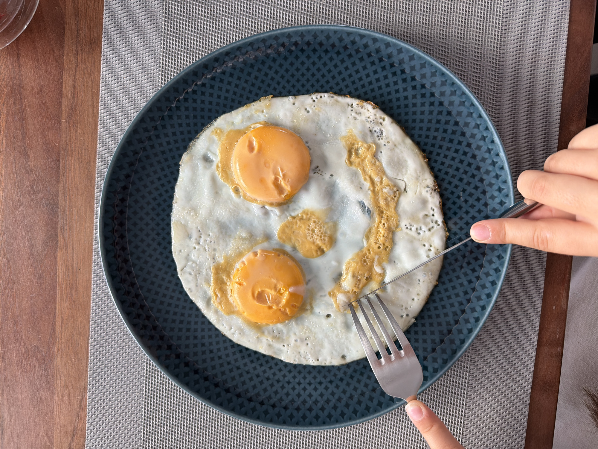 A close-up, overhead shot shows a fried egg on a dark blue plate, arranged to resemble a smiley face. The two egg yolks form the eyes, and the fried egg white, with browned edges, outlines the face. A fork and knife are held by a person's hands, with the fork positioned near the edge of the egg white, suggesting preparation for eating. The plate rests on a grey placemat, which is on a wooden table. The scene is domestic, likely a breakfast setting. This image is not set in Poing, Germany.