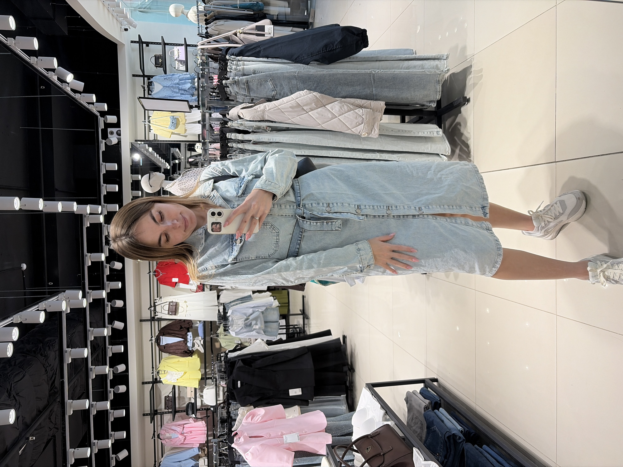 A woman is taking a selfie in what appears to be a clothing store. She is wearing a light blue denim dress and white sneakers. Her hair is long and light brown. She is holding a white smartphone. Behind her, clothing racks are filled with various garments, including jeans, jackets, dresses, and shirts in different colors. There is an array of lighting fixtures on the ceiling. The floor is light-colored tile. The location may be in Izhevsk, Russia, but there is no explicit indication of the city in the image.