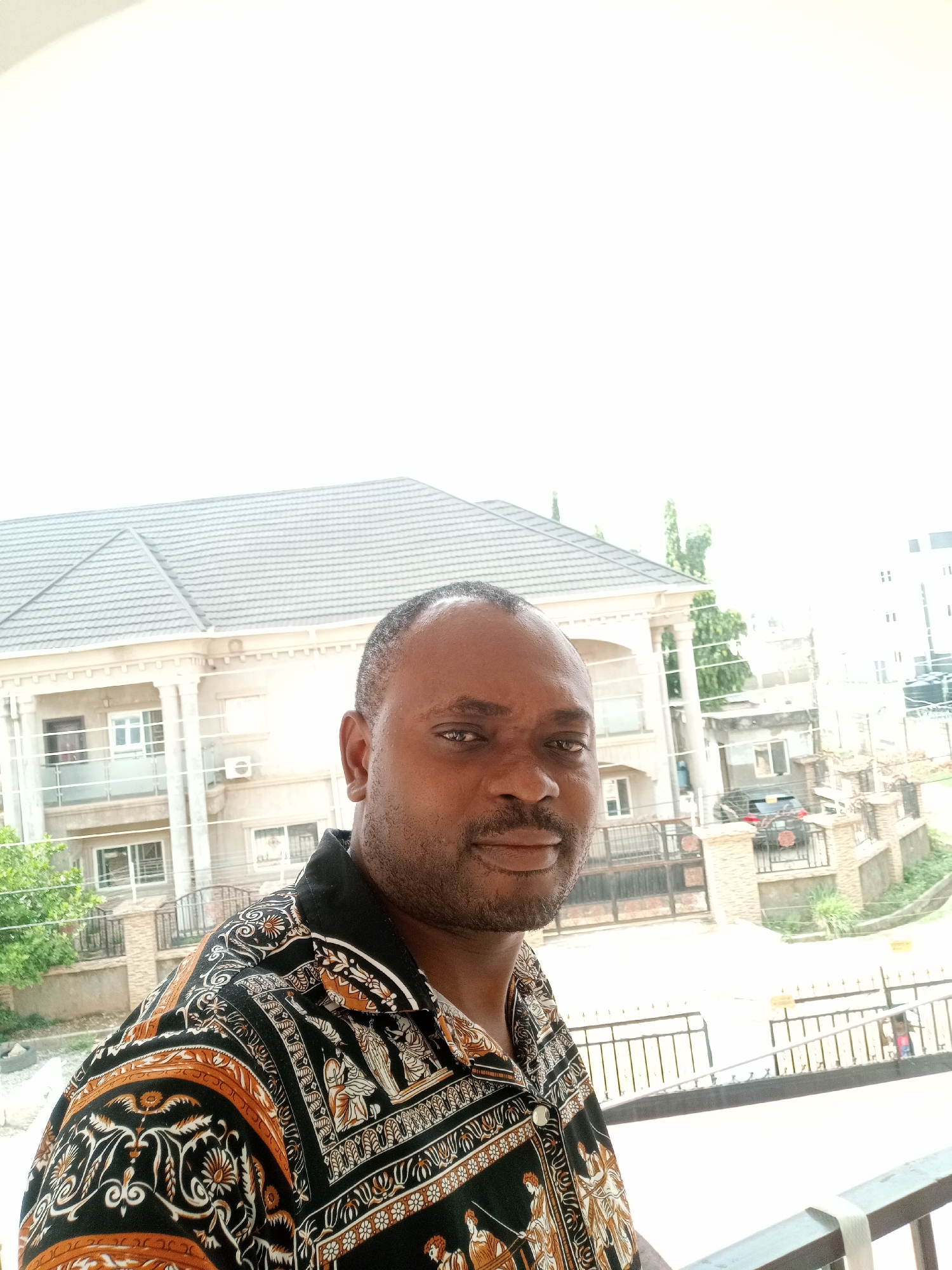 A medium shot depicts a man, likely in his late 30s or early 40s, with short dark hair and a clean-shaven face, facing the camera. He is wearing a patterned shirt with a black background and ornate designs in orange and white.
In the background is a two-story building, possibly a residential house with a grey tiled roof, multiple windows, and a beige exterior. A car is visible behind a gated entrance to the property. Fencing surrounds the property. A neighboring building is also visible in the distance.
The location appears to be an outdoor area, potentially a balcony or raised platform, within Gwarinpa, Nigeria.