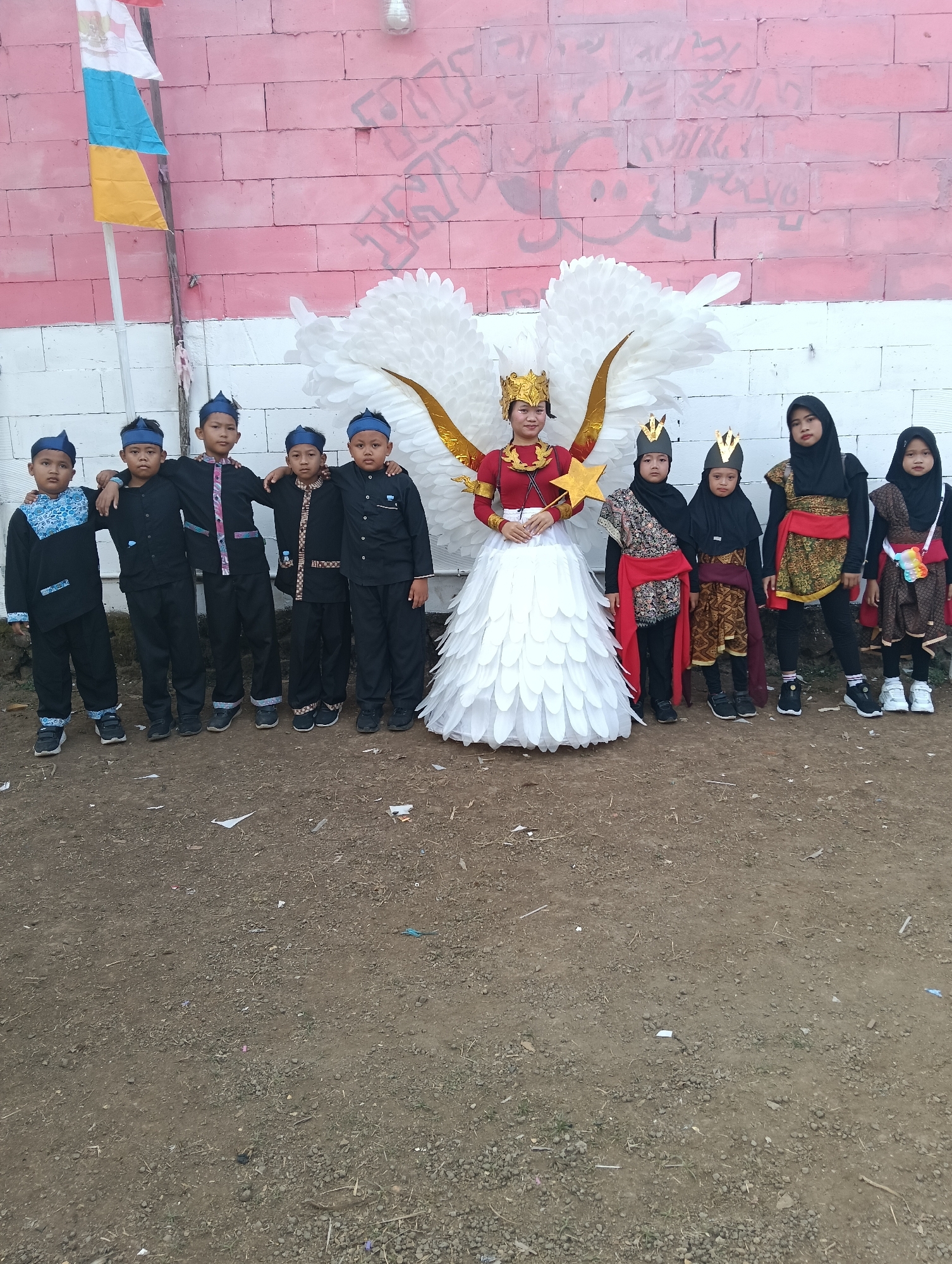 In Tasikmalaya, Indonesia, a group of children poses for a photograph in front of a pink and white brick wall. The wall is adorned with graffiti. To the left of the group, a flag with white, blue, and yellow sections is mounted on a pole.
At the center of the image is a young person dressed in a striking costume. They wear a golden crown, a red top, and a voluminous white skirt made of layered, petal-like material. Large, white wings, some with golden accents, extend from their back.
To the left of this central figure are five young boys. They are dressed in matching black outfits consisting of long-sleeved shirts and pants, each adorned with different patterned trim. They wear blue headbands.
To the right of the central figure are four young girls. They are all wearing headscarves. Three of them wear dark headscarves, and one wears a black headscarf. Their outfits vary, with some wearing traditional patterned fabrics, red sashes, and elaborate vests. One of the girls on the far right wears a multi-colored accessory over her shoulder.
The ground in front of the group is composed of dry dirt, gravel, and scattered pieces of paper and debris. The lighting suggests it is daytime.