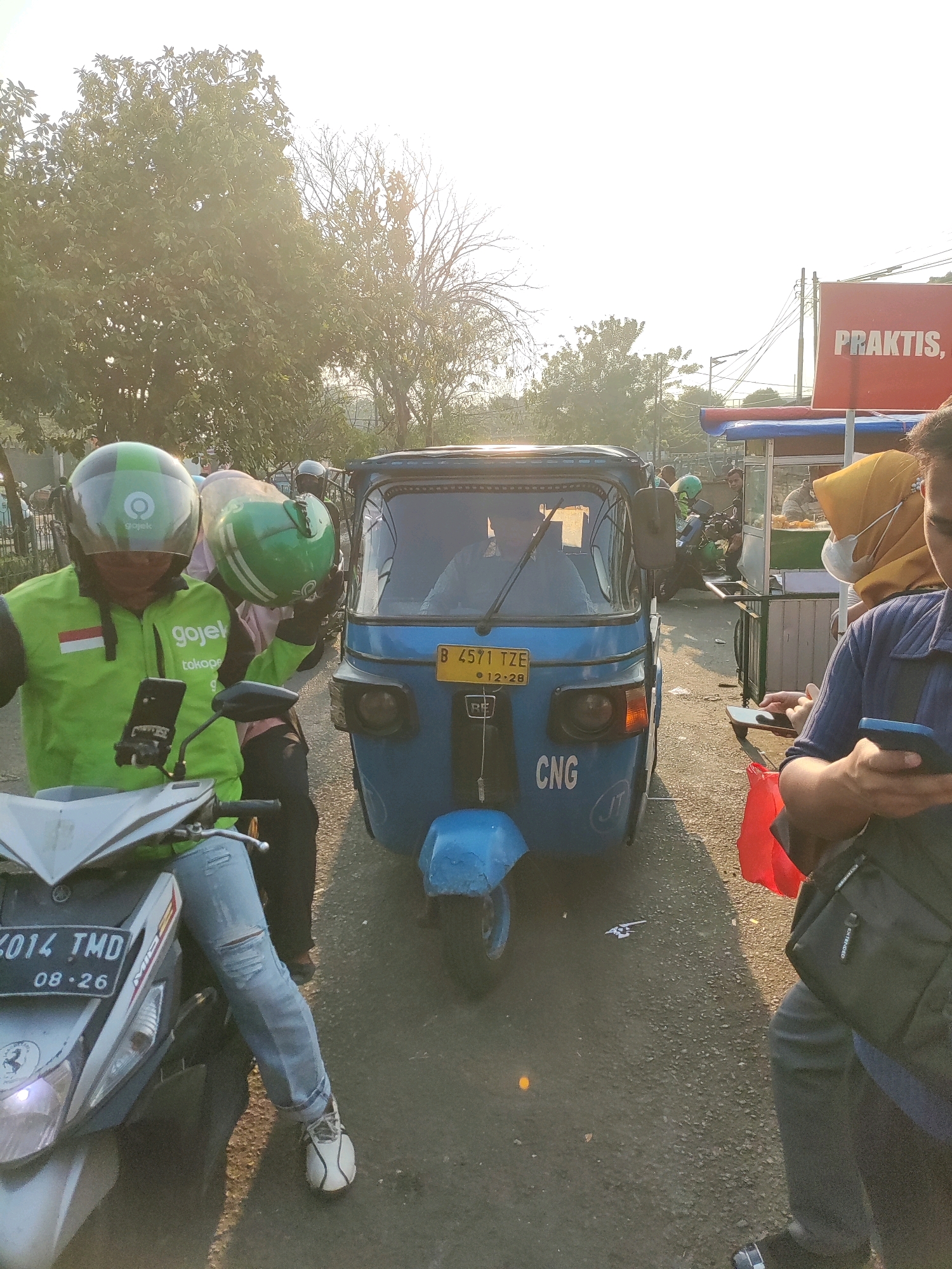 The image captures a street scene in East Jakarta, Indonesia, with a bright, hazy sun suggesting late afternoon. In the foreground, a person wearing a bright green Gojek jacket and helmet is seated on a silver motorcycle, holding a phone mounted on the handlebars. To their right, a blue three-wheeled vehicle, likely an auto-rickshaw, is prominently featured. The auto-rickshaw displays a yellow license plate with the text "B 4571 TZE" and "12-28". The words "CNG" are visible on the rear of the auto-rickshaw, indicating it runs on compressed natural gas. Further back, other vehicles and people are visible, including a food cart with a vendor and customers, and a person in a yellow hijab and mask. A red sign with the word "PRAKTIS" is also visible in the background. The environment appears to be a typical urban street with trees lining the background and pavement underfoot. The overall impression is a snapshot of daily life and transportation in a bustling Indonesian city.