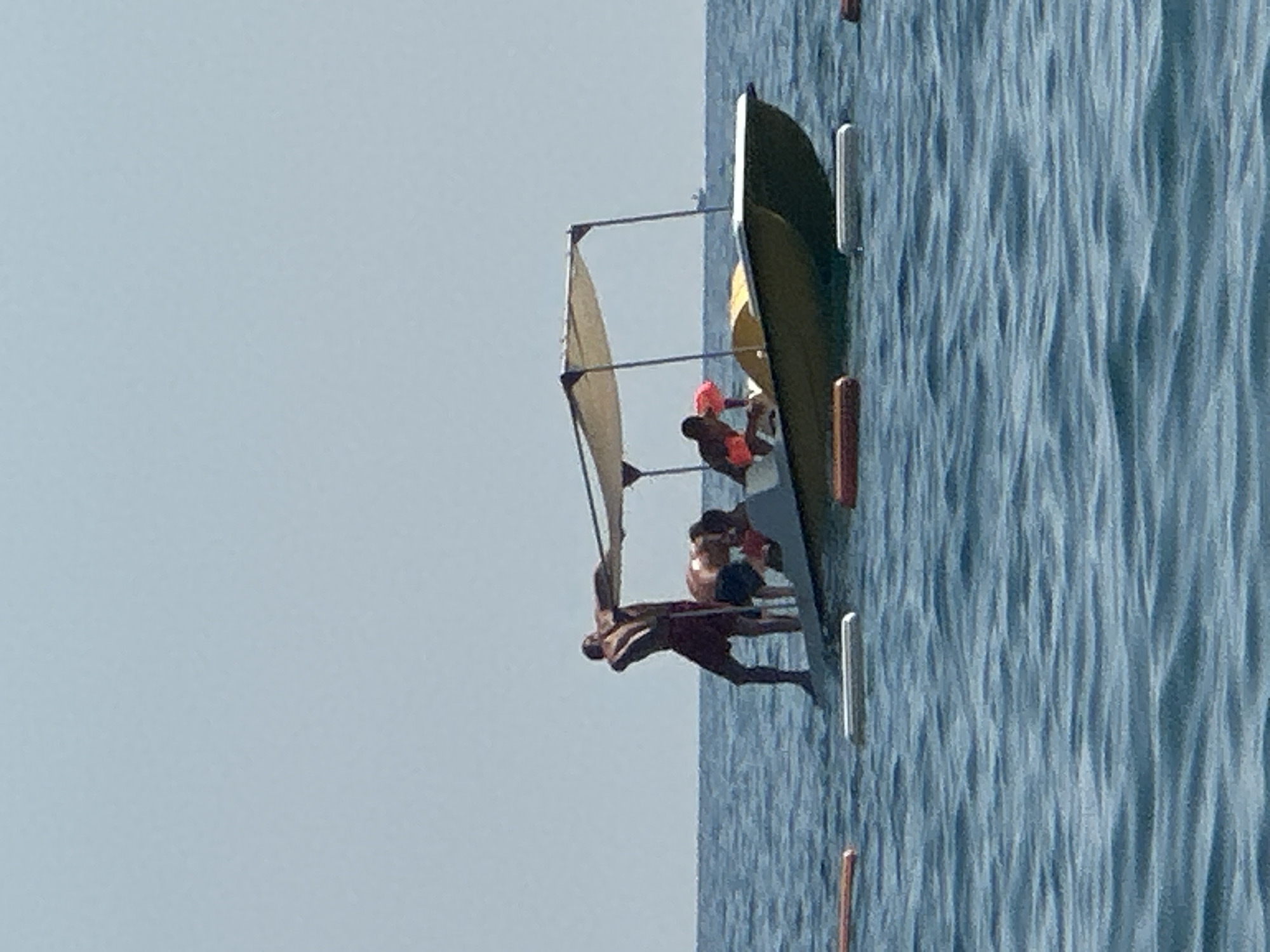 A person stands on a paddle boat with a canopy, while another person bends down on the boat. In the background, a vast expanse of calm blue water meets a clear, pale blue sky. The paddle boat is yellow and gray. The people on the boat are wearing swim trunks. There are several orange and white buoys in the water in front of the boat.