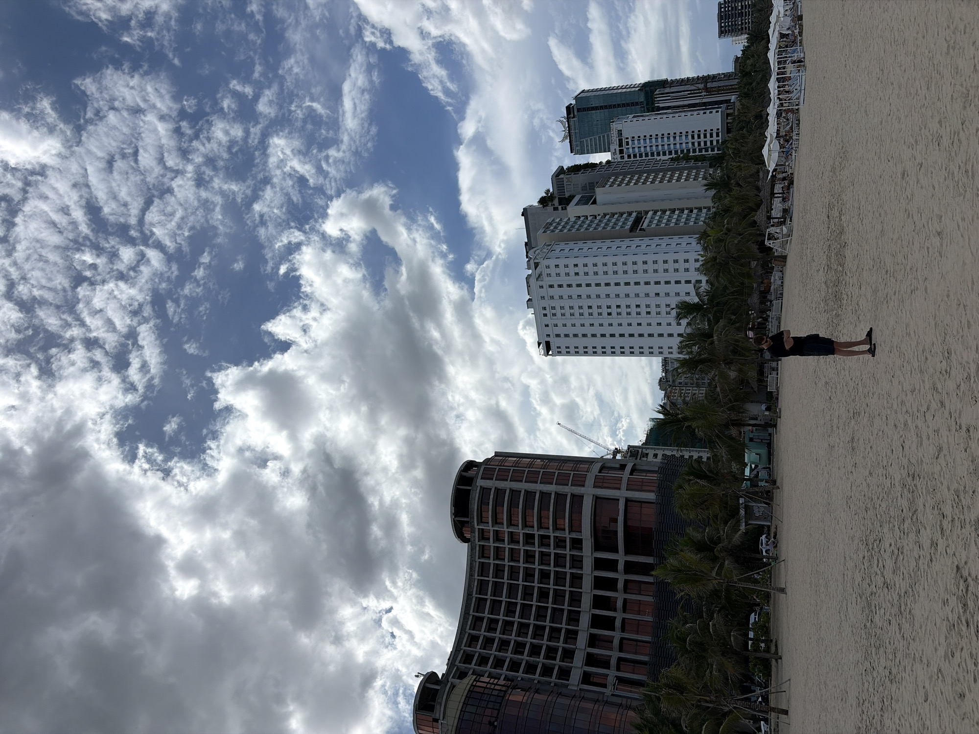 A man stands on a sandy beach in the city of Đà Nẵng, Vietnam, looking out towards the ocean. He is wearing a dark t-shirt and blue shorts, with his arms crossed. The beach is lined with palm trees and rows of white umbrellas, with lounge chairs underneath. In the background, several modern high-rise buildings stand tall against a cloudy sky. One building, on the left, has a curved facade and many windows. Another building, in the center, is white with many small windows. In the distance, construction cranes are visible, indicating ongoing development. The sky is a mix of white clouds and patches of blue.