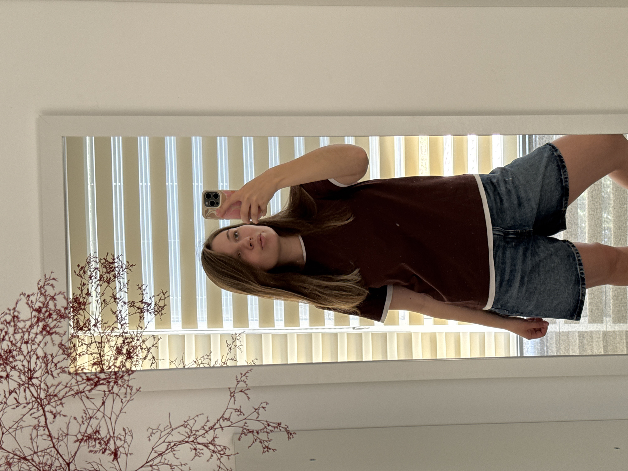 In the city of Budva, Montenegro, a young woman is depicted taking a mirror selfie. She has long, straight brown hair, fair skin, and is wearing a dark maroon t-shirt with white trim on the collar and sleeves, and blue denim shorts. Her right arm is raised, holding a smartphone with a pink and white swirled protective case, positioned to capture her reflection. Her lips are slightly pursed, and her gaze is directed towards the phone/mirror. The reflection shows a room with a large window covered by light-colored horizontal blinds, allowing ambient light to enter. To the left of the mirror, a decorative plant with thin, reddish-brown, branched stems is visible against a white wall. The mirror itself has a simple white frame, and plain white walls flank it on both sides.