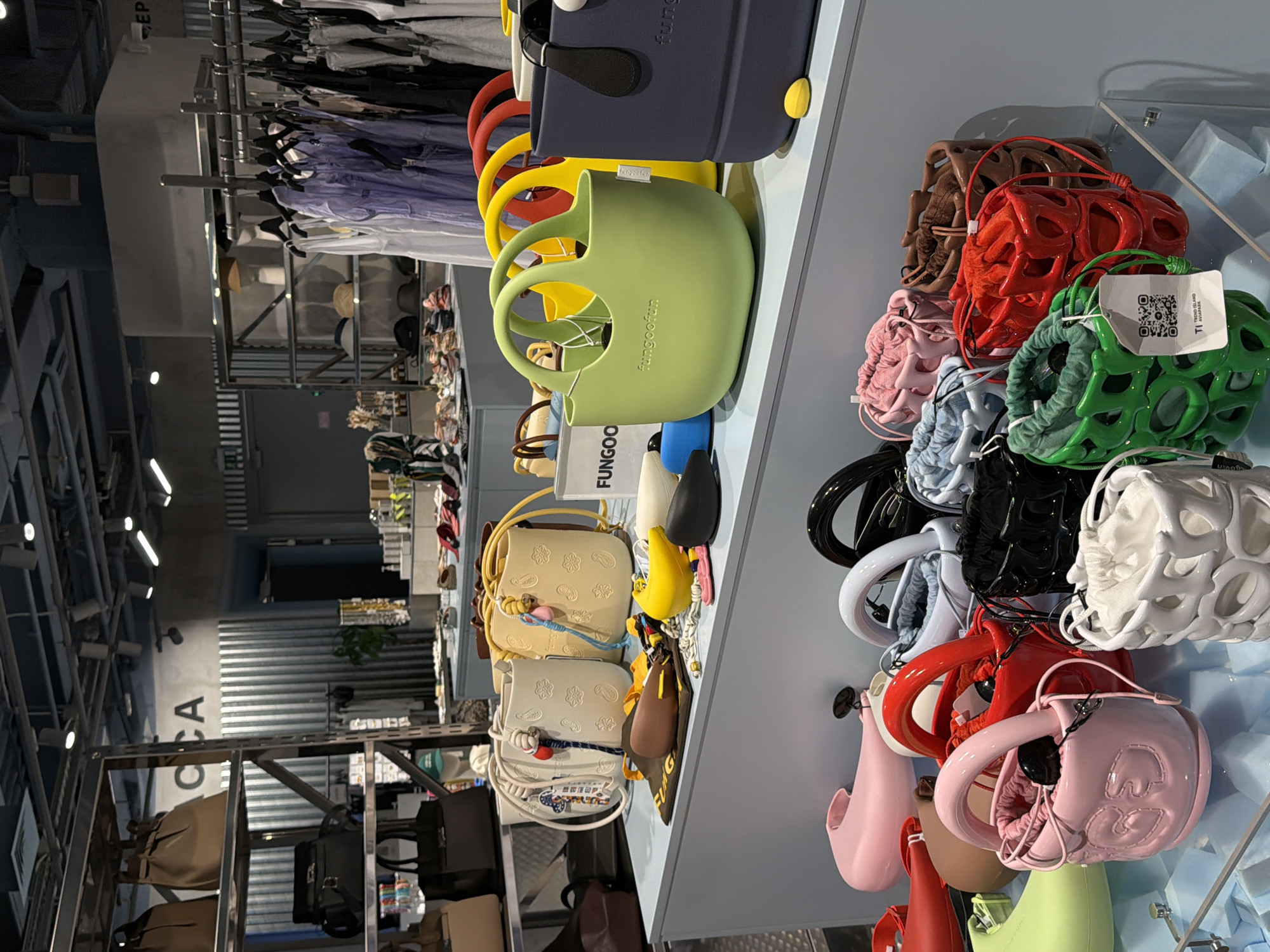 This media file captures a well-lit interior scene, likely a retail store or boutique, showcasing a vibrant display of handbags and accessories. The overall impression is one of organized merchandising, with items neatly arranged on shelves and hangers.
The primary subjects are the various handbags, which come in a wide range of colors and styles. Several are small, bucket-style bags, some with intricate patterns and embellishments like embossed flowers or beaded drawstrings. Others are larger tote bags in neutral tones. A prominent feature is a row of brightly colored, rubber-like bags with rounded shapes and distinct handles, including lime green, yellow, and red. There are also more structured handbags in black and white with unique, cage-like designs. The items appear to be new and well-maintained, suggesting a retail environment.
In the background, clothing items are visible hanging on racks, including what appear to be t-shirts and sweaters in various colors, from pastels to darker hues. This reinforces the idea of a clothing or accessory store. The setting is modern, with sleek shelving units and visible lighting fixtures. The light is bright and even, suggesting it is daytime or the store is well-illuminated.
There are no people visible in the frame, so no human activity is depicted. However, the arrangement of the merchandise implies a commercial setting where customers would browse and purchase items.
Notable details include the specific brands or labels on some items. One tag on a green bag clearly displays a QR code and the text "TRESI ISLAND AVAPARK". Another tag on a dark blue bag reads "fungo". The arrangement of the colorful rubber bags, with their bold colors and unique forms, is a striking visual element. The overall mood is clean, organized, and visually appealing, aiming to attract shoppers. The location context is Moscow, Russia, which might inform the specific brands or styles present.