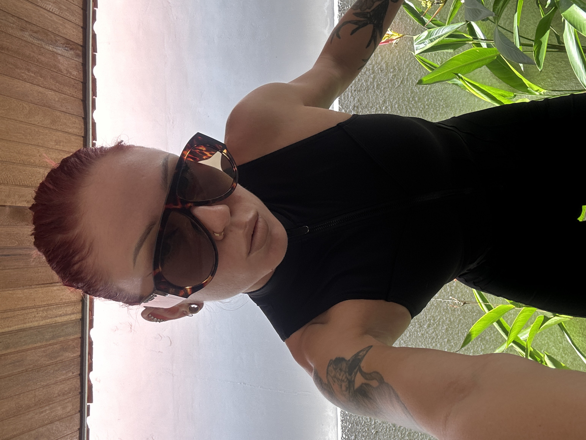 A woman with reddish-brown hair and tattoos on her arms is wearing sunglasses and a black outfit. She appears to be outdoors, near water and lush greenery, possibly in a tropical setting in North Kuta, Indonesia. The background suggests a combination of natural and constructed elements.