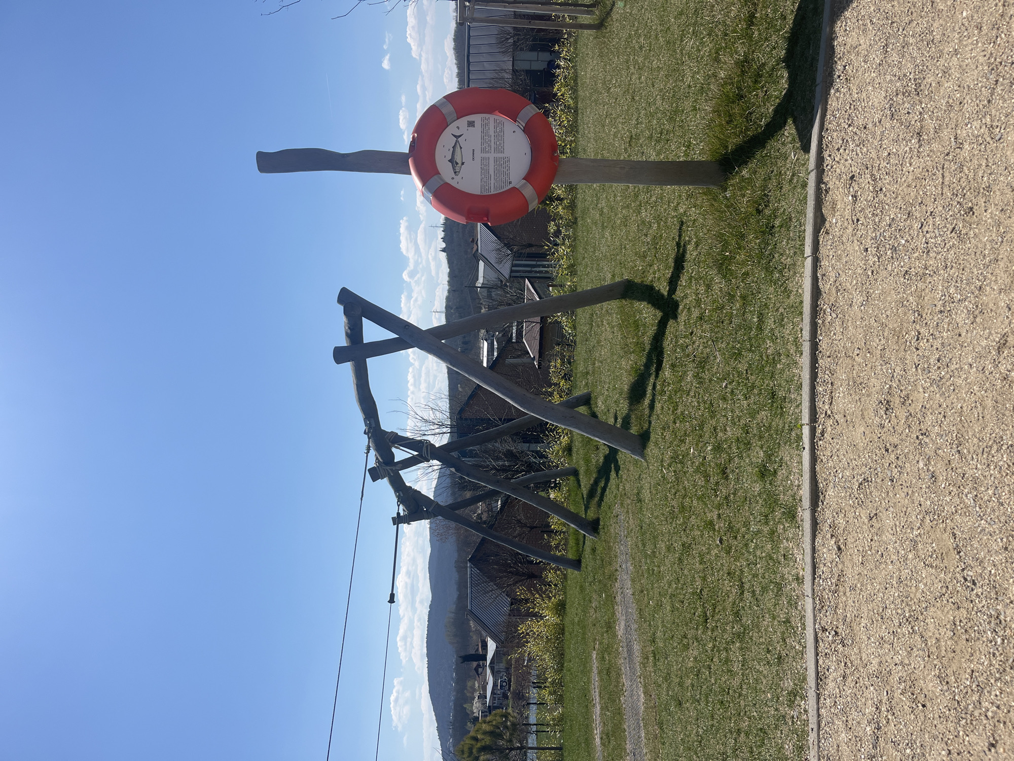 The image depicts an outdoor scene in Burgas, Bulgaria, featuring a small wooden zip line structure. The zip line has two wooden support towers connected by a cable. A wooden post with a circular red and white life preserver attached stands nearby; the life preserver has an informational sign with a fish illustration. The ground is covered with green grass, adjacent to a gravel path. Buildings are visible in the background, and the sky is clear blue with scattered clouds.