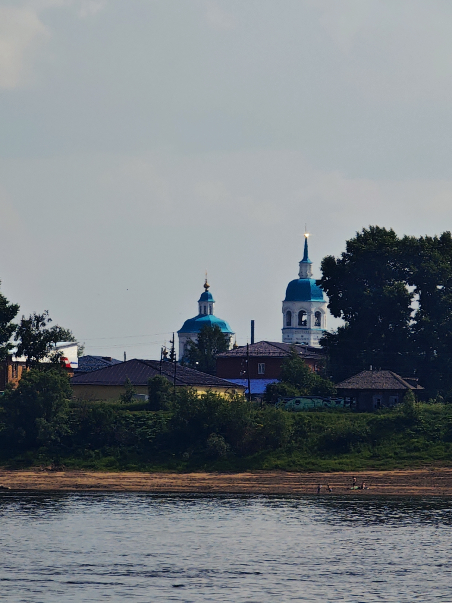 The image captures a view of the Yeniseysk Monastery in Yeniseysk, Russia, from across the Yenisei River. The monastery's distinct blue-domed churches and a tall bell tower are visible above a line of trees and houses situated on the riverbank. The water in the foreground reflects the overcast sky. Several small figures are present on the sandy shore, suggesting leisure activity. The overall scene depicts a tranquil, semi-rural setting with religious architecture dominating the skyline.