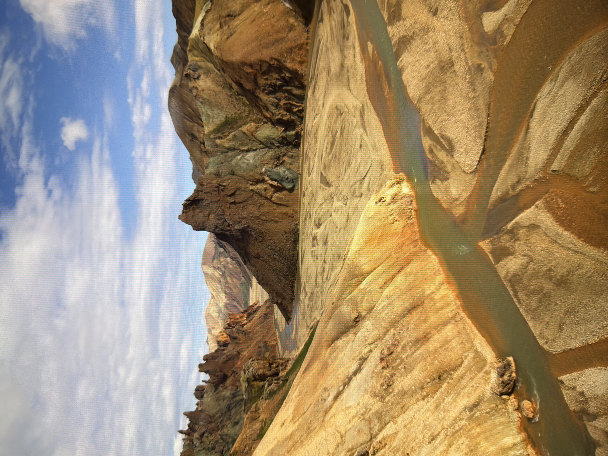 The image depicts a dramatic and colorful landscape of rhyolite mountains and a braided river. The mountains exhibit a range of earthy tones, from deep browns and reds to ochre and yellow, with streaks of green and grey in some areas, suggesting mineral deposits. The terrain is rugged and eroded, with sharp peaks and steep slopes. A wide, shallow river snakes through the valley, its waters a greenish-grey. The riverbed is composed of sand and gravel, with intricate patterns formed by the flowing water. The sky above is a mix of blue and white, with scattered clouds. The overall impression is one of a remote and wild natural environment. The image does not contain any elements that would suggest it is located in Kyiv, Ukraine.