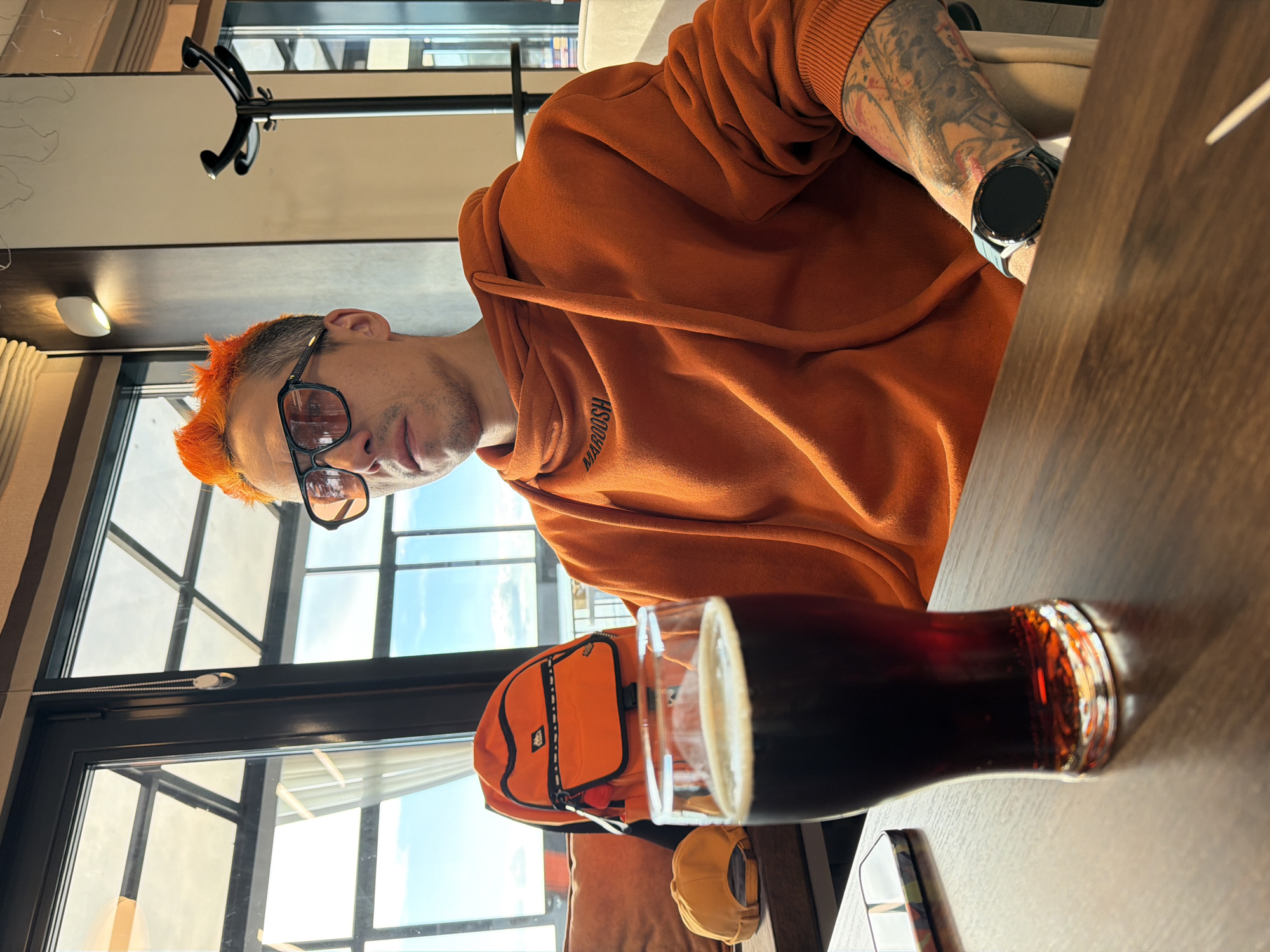 A man with distinctive orange, spiky hair is seated at a wooden table in what appears to be an indoor establishment, likely a cafe or bar, located in Chaikovsky, Russia. He is looking directly at the camera with a neutral expression, wearing sunglasses and light stubble. He is dressed in an orange hooded sweatshirt with the word "MAROOSH" embroidered on the front. His left arm is visibly tattooed, and he wears a dark smartwatch with a blue-grey strap.
In front of him on the table is a glass containing a dark beverage, likely beer, with a light head. An orange backpack or small bag is also on the table, consistent with the man's attire color. A yellow baseball cap is partially visible next to the bag.
The setting is brightly lit by natural light streaming through large windows in the background, revealing a clear blue sky and the exterior of a modern building. Inside, a black coat rack is visible, along with a wall-mounted light fixture. The scene depicts a casual daytime moment.