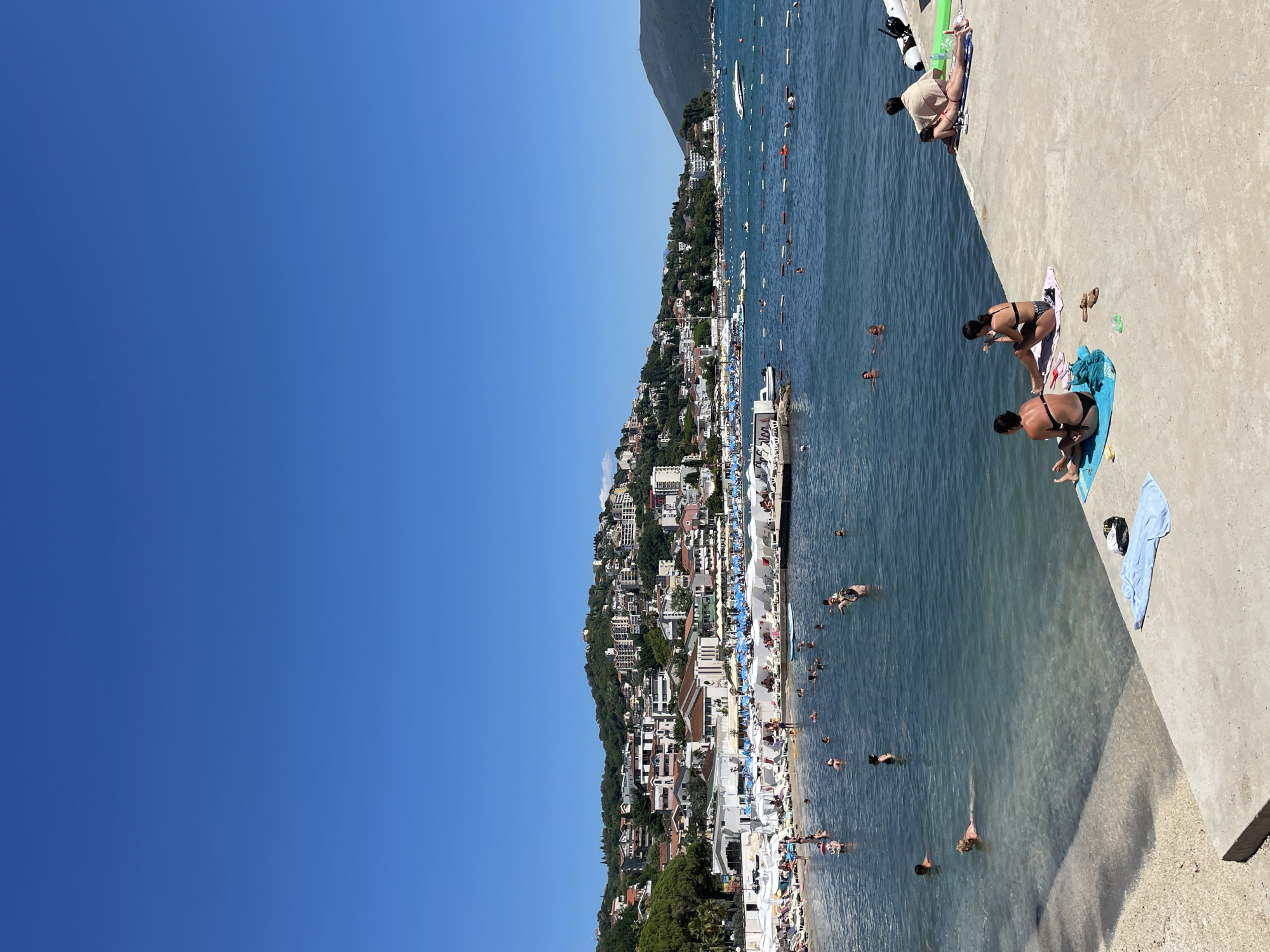 In Igalo, Montenegro, a crowded beach scene unfolds under a clear blue sky. The foreground features a concrete pier where several individuals are seen relaxing. To the left, two women in bikinis are seated on towels, one appearing to eat and the other looking away. Further along the pier, a person in a light-colored cover-up is lying down, facing away from the camera, while another person is positioned on their stomach. The water is filled with people swimming and wading, enjoying the sunny day. Beyond the water, a bustling beach is lined with numerous white umbrellas and lounge chairs, indicating a popular tourist destination. A hillside covered with buildings and lush greenery forms the backdrop, characteristic of coastal towns in the region. Boats are visible scattered across the water, and the shoreline stretches towards a distant mountain range.
