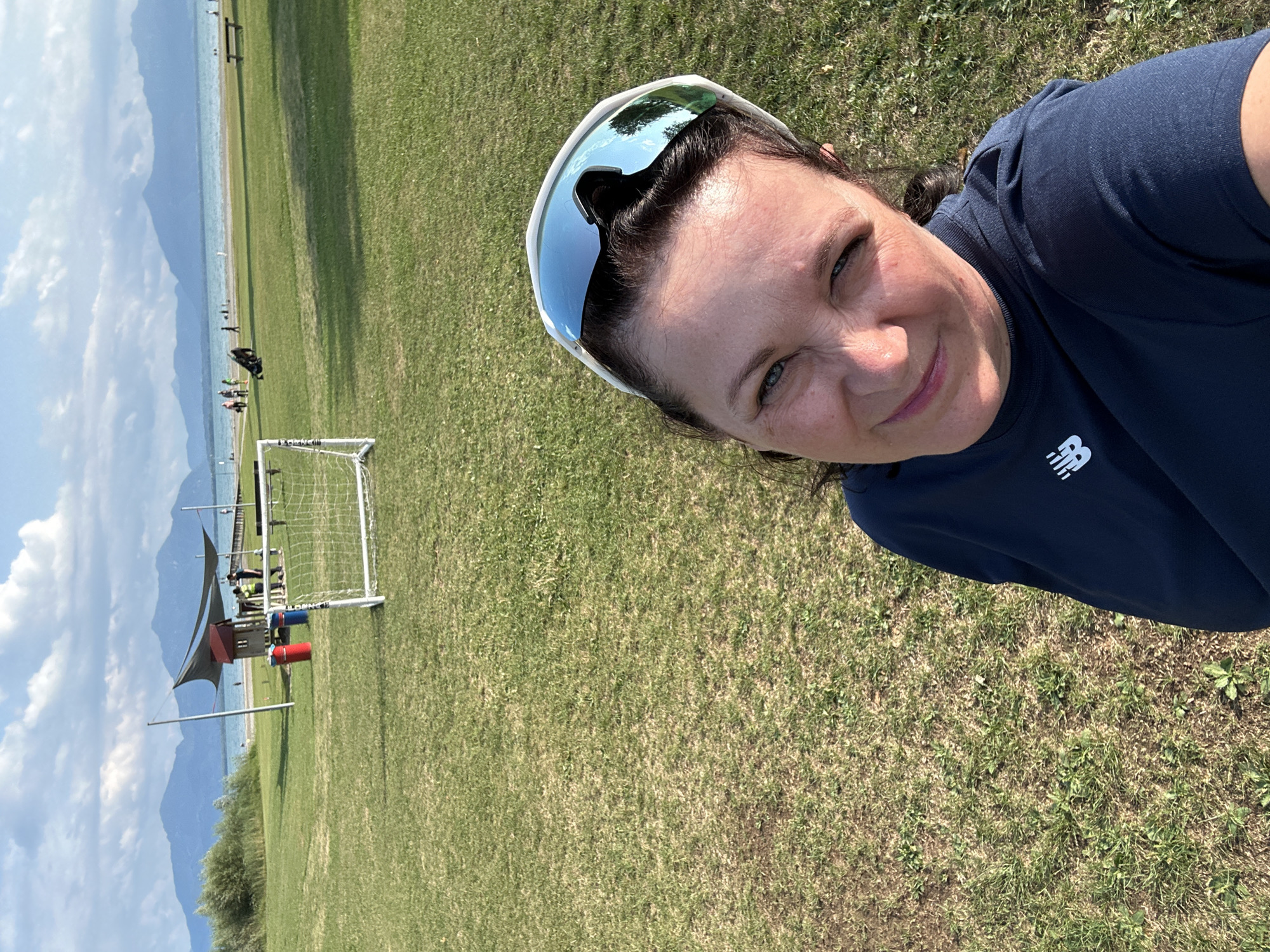 A woman is taking a selfie in a grassy park near a lake. The woman is wearing a dark blue shirt and white sports glasses. In the background, there is a small soccer goal, a playground structure with two trash cans, a pier, a beach, a body of water, and mountains in the distance. The sky is partly cloudy. The location is identified as Seeon-Seebruck, Germany.