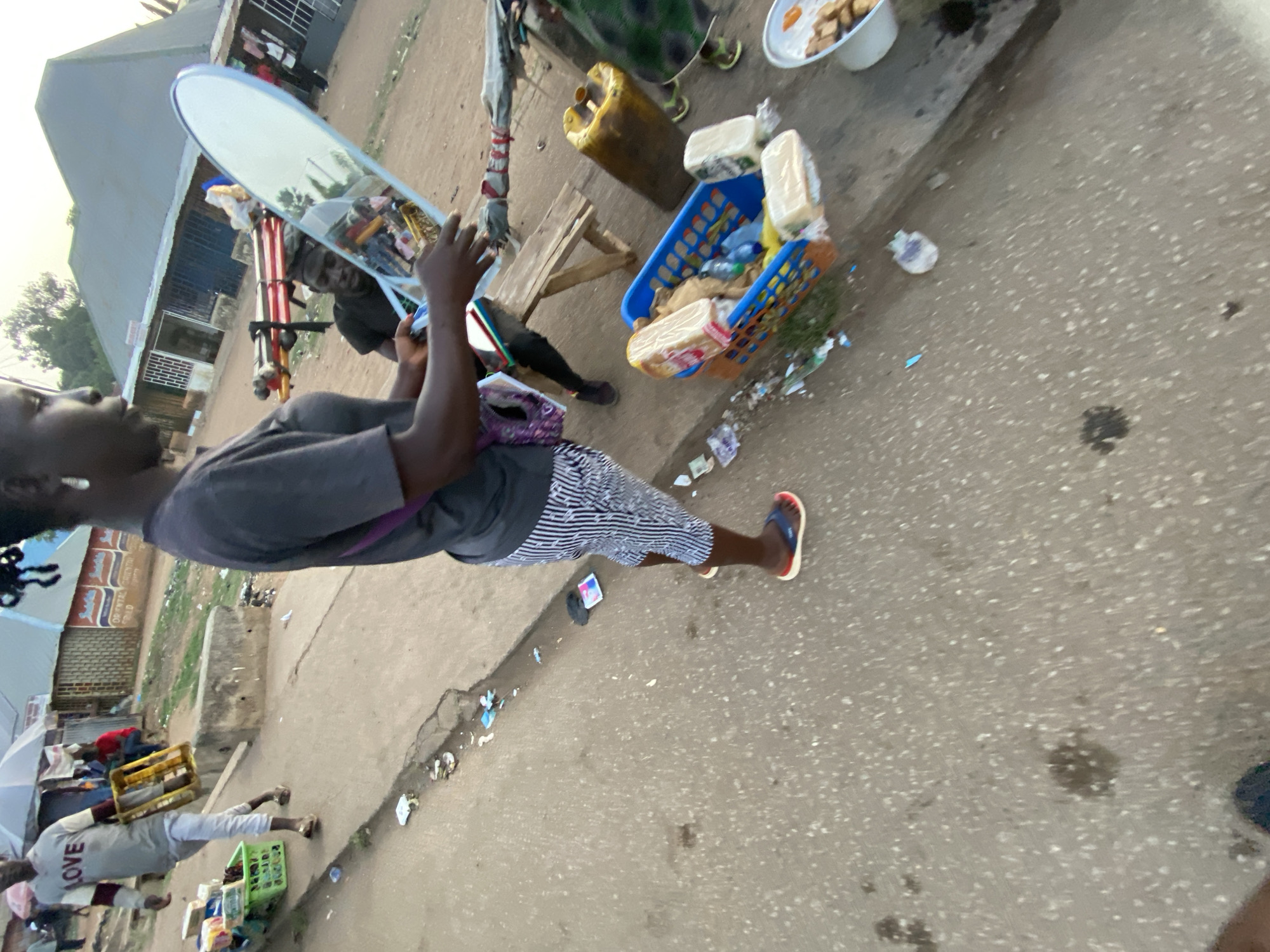 This street-level image, captured in Bukuru, Nigeria, depicts an everyday scene of an informal market or sidewalk activity. The photo is rotated, showing subjects and objects at an angle.
In the immediate foreground, a person with dark skin is visible from the chest up, wearing a grey t-shirt and striped shorts. A purple bag strap crosses their shoulder. They are holding a large, oval-shaped mirror with both hands. Reflected in the mirror is another person with dark skin, wearing a dark t-shirt, presumably a vendor or another individual present. The foreground person is also wearing red and blue flip-flops.
To the right of the main subject, various goods are laid out on the ground and in containers. A blue plastic basket contains several packaged loaves of bread or similar goods, along with plastic bottles. Nearby, two white bowls hold what appear to be cooked food items, possibly snacks, on a small wooden stool. A yellow jerrycan is also present. The ground is paved, littered with small pieces of paper and plastic.
Further down the street to the left, another individual is seen wearing a light grey t-shirt with the word "LOVE" written across the chest, and white pants, carrying a yellow crate on their shoulder. Behind them are glimpses of market stalls and a white umbrella. Buildings with corrugated metal roofs and concrete walls line the street in the background. On one building, faint text reads "DR. OPTICAL DENTAL HOSPITAL".
The lighting suggests late afternoon or early morning, with a soft, warm glow. The overall scene captures the bustling, informal commerce typical of a Nigerian street market.