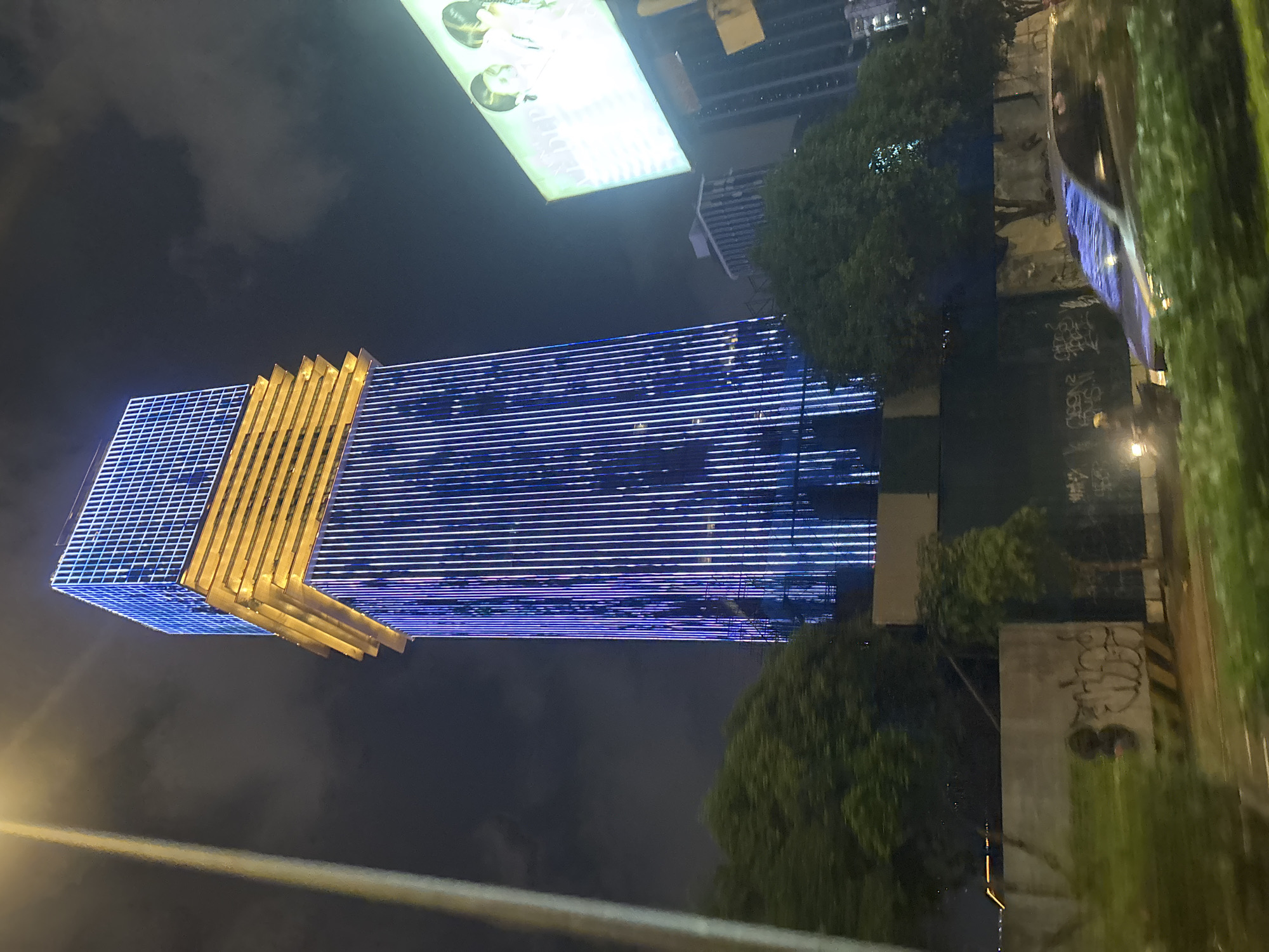 A vertical, low-light photograph shows a modern skyscraper illuminated with blue and white lights at night. The building has a unique architectural design with a grid-like pattern on one section and horizontal blue lights spanning most of its facade. A golden, stepped section is visible on the left side of the building. Trees, also dark and silhouetted, are scattered around the base of the skyscraper. In the background, other buildings are visible, some with lights on. A large, brightly lit rectangular sign with images of people is positioned above and to the left of the skyscraper. The overall scene suggests an urban environment at night. The image is slightly tilted, and there are some blurry green elements, possibly foliage or a reflective surface, in the foreground on the right. The sky is dark and cloudy. No specific emotions are conveyed.