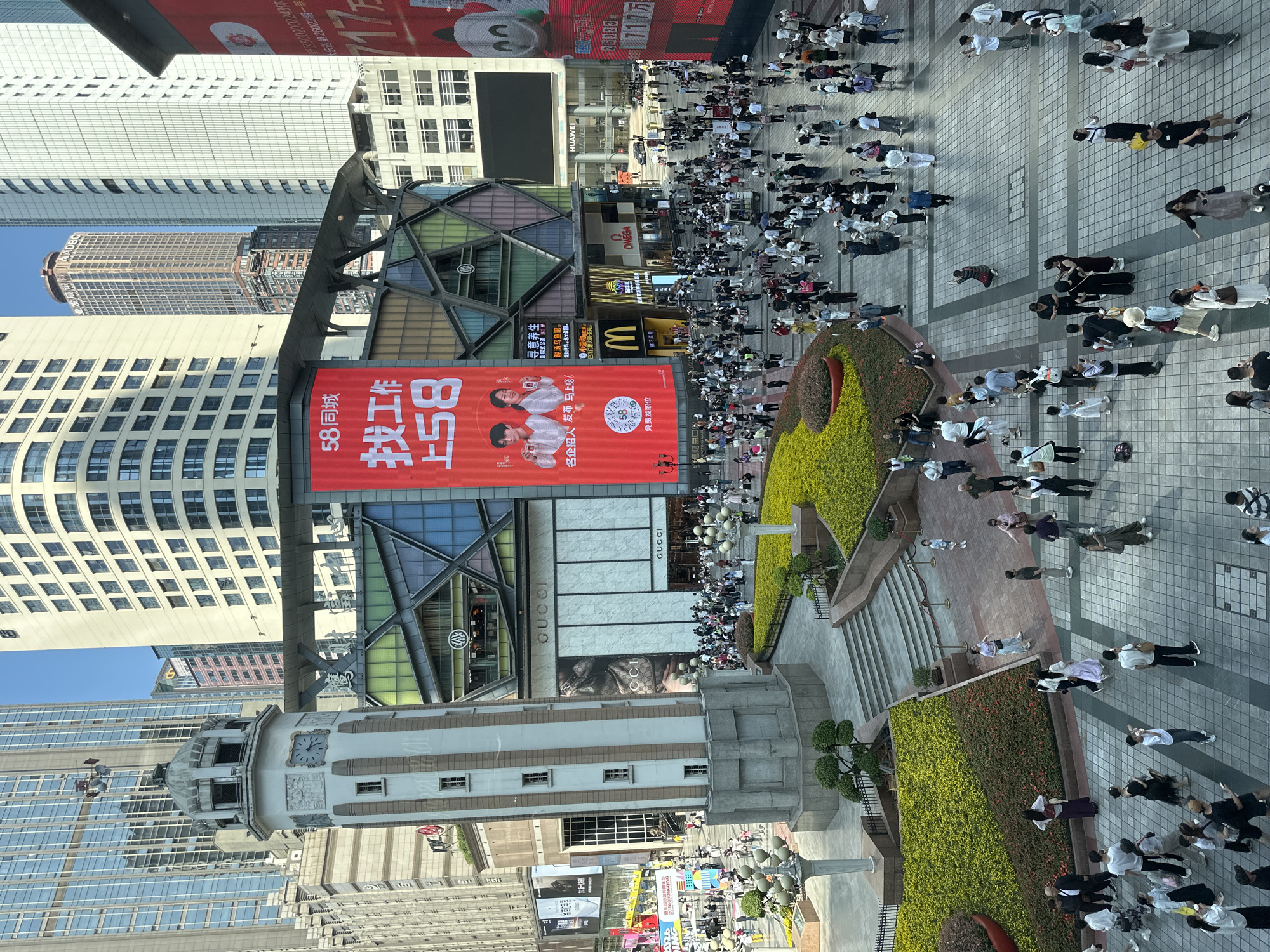 This elevated wide shot captures a bustling commercial district in Yuzhong District, China, under a clear blue sky. A large crowd of people, dressed in casual attire, fills a spacious paved square, walking in various directions. Many appear to be looking at their phones or interacting with others.
The scene is dominated by a mix of modern and contemporary architecture, including several towering skyscrapers with reflective glass and patterned facades. In the foreground, a distinctive white and gray clock tower stands prominently, featuring a circular clock face. Planters with lush green and yellow foliage line the walkways and steps, adding natural elements to the urban environment.
Numerous commercial buildings and storefronts are visible, displaying well-known brand logos such as GUCCI, McDonald's, OMEGA, and HUAWEI. A large red digital billboard is a focal point, displaying Chinese characters that prominently feature "58同城" (58.com), "找工作" (find a job), and the number "58". Smaller text on the billboard indicates "名企招人发布 马上发" (Famous enterprises recruiting, publish immediately) and "免费发职位" (post jobs for free), alongside images of two young individuals holding smartphones. Other discernible Chinese text includes "寻意养生", "影院式足道246", "酸汤乌鱼馆", and "小荣和".
The bright daylight casts strong shadows across the square, indicating a sunny day. The overall impression is one of a vibrant, active urban center during daytime hours.