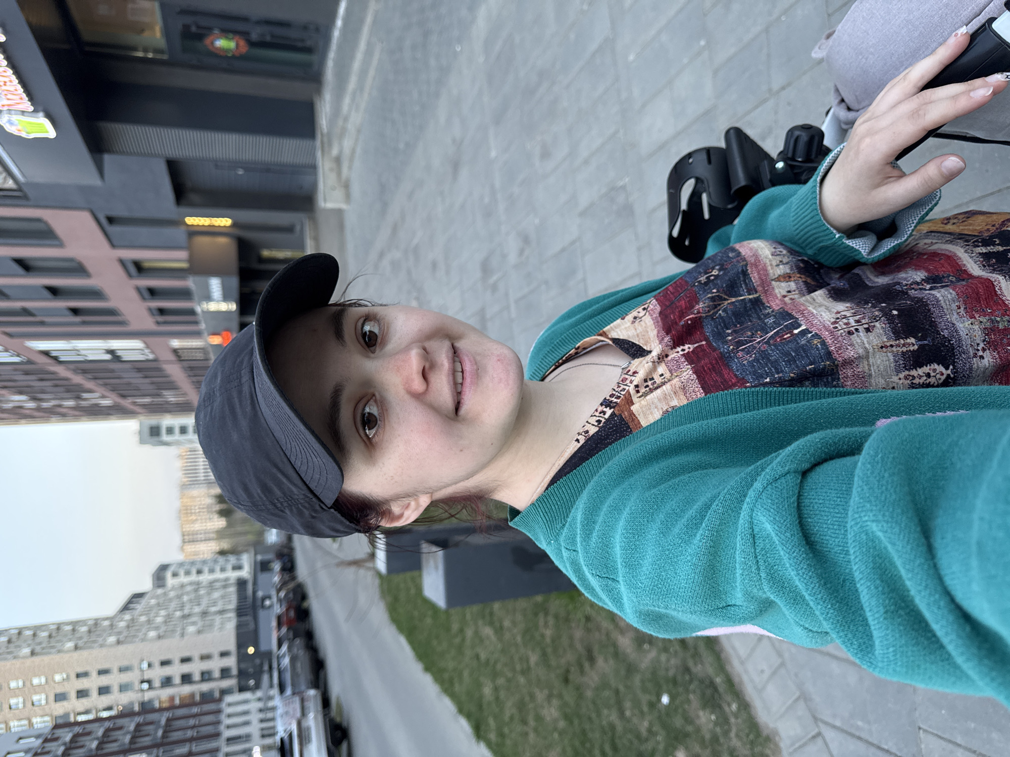 A young woman wearing a black cap and a green sweater over a patterned dress is taking a selfie outdoors. She is holding the handle of a stroller. The location is Izhevsk, Russia, as evidenced by the buildings in the background. The buildings appear to be modern residential or commercial structures. There's a sidewalk and some green space visible to the right of the woman. Cars and other buildings line the street behind the woman. The sky is overcast.