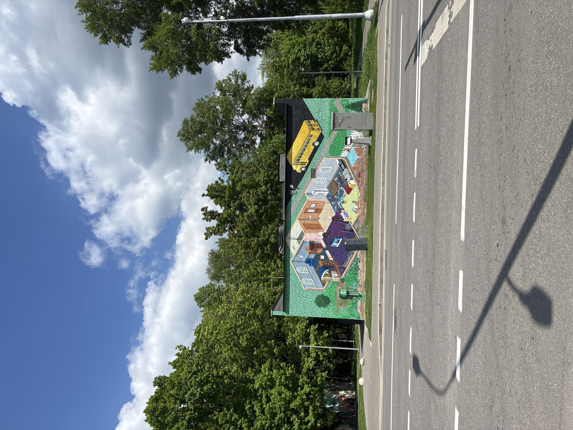 The image depicts a building with a colorful mural located on a street in Moscow, Russia. The mural features a yellow school bus, houses, and other objects. The sky above is partly cloudy with blue patches. Trees surround the building. A road with white lane markings is visible. A lamppost casts a shadow on the road.
