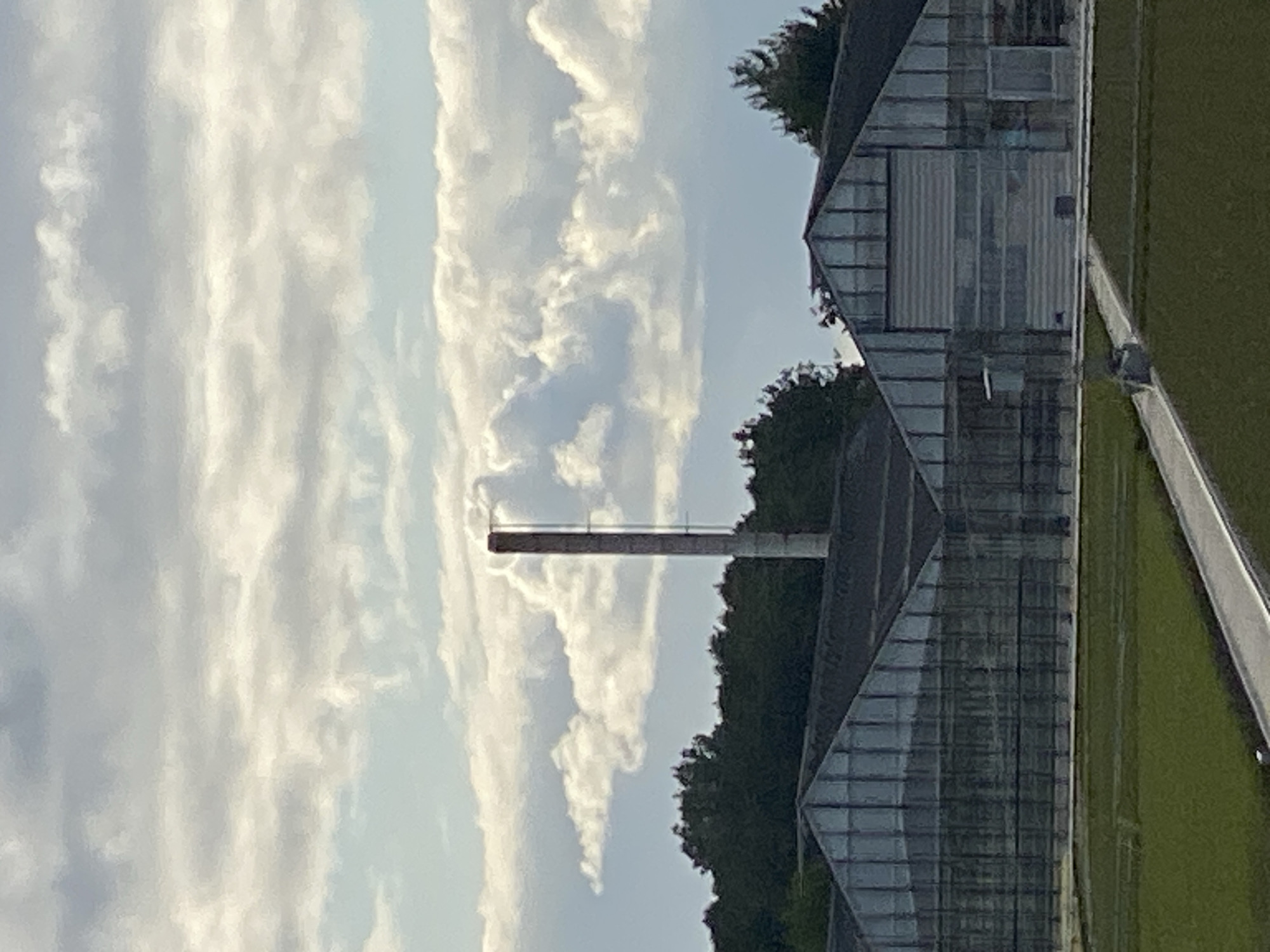 An outdoor, mid-range shot captures a cloudy sky over a field and greenhouses. The sky is filled with dramatic, layered clouds in shades of gray and white, with patches of blue visible. A tall, slender, concrete smokestack stands prominently in the midground, positioned slightly to the right of the frame. In the foreground and extending across the lower portion of the image are several large greenhouses with glass roofs and walls, suggesting an agricultural or industrial setting. Dark green trees form a dense line behind the greenhouses, separating them from the sky. A manicured green field is visible in front of the greenhouses, with a light-colored pathway running through it. The overall impression is one of a functional, possibly industrial, area under a dynamic sky. The location of Schwarme, Germany is not visible within the image itself.