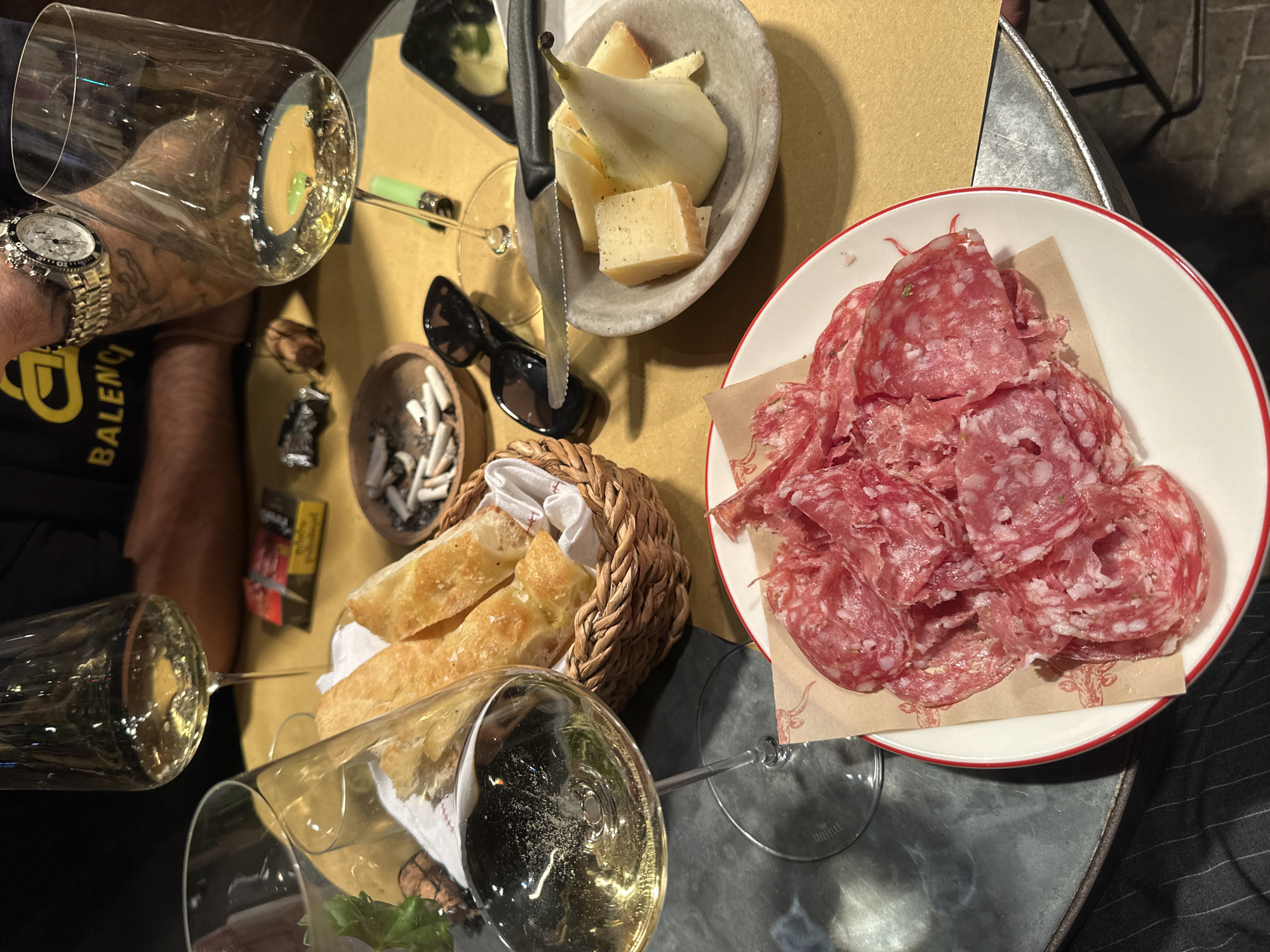 The image shows a high-angle view of a table laden with food and drinks, suggesting a gathering or meal. On the table, there are two wine glasses filled with a light-colored liquid, possibly white wine. A plate of sliced salami, a bowl containing cheese and slices of pear, and a basket of bread are also present. The scene is likely indoors, given the controlled lighting. A person's arm, wearing a watch, and clothing with the word "BALENCI" are visible near the glasses. There's an ashtray containing cigarette butts and a pair of sunglasses on the table. The food and drink arrangement indicate a charcuterie board. The environment seems upscale or casual dining. There is no indication of the location being in London, United Kingdom.