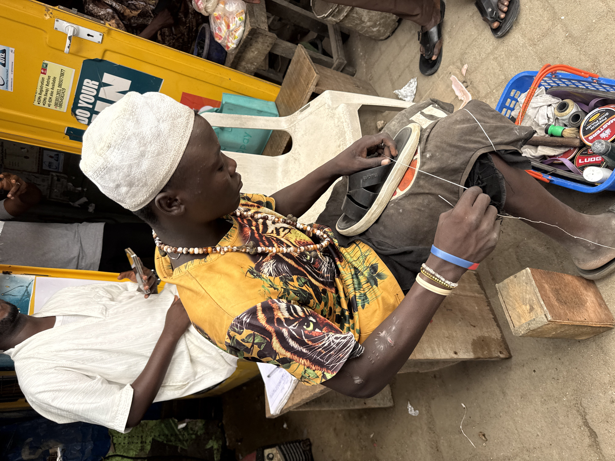 In a bustling outdoor market stall in Lekki, Nigeria, a young man is engrossed in repairing a black sandal. He is seated on a wooden stool, wearing a white embroidered cap, a bright yellow t-shirt with a tiger print, and several beaded necklaces. His arms are adorned with bracelets, including a blue and red one. He is meticulously stitching the sandal with white thread, his focus evident as he works. Nearby, a blue plastic basket overflows with shoemaking supplies, including cans of shoe polish, brushes, and spools of thread. To his left, a wooden crate holds a notepad and pen. In the background, a yellow kiosk stands tall, featuring various advertisements and a handle, suggesting it might be a service point. Another person, partially visible, wears a white traditional garment. The ground beneath is a mix of concrete and dirt, typical of an open-air market. The scene is dynamic, with elements of commerce and skilled craftsmanship visible.